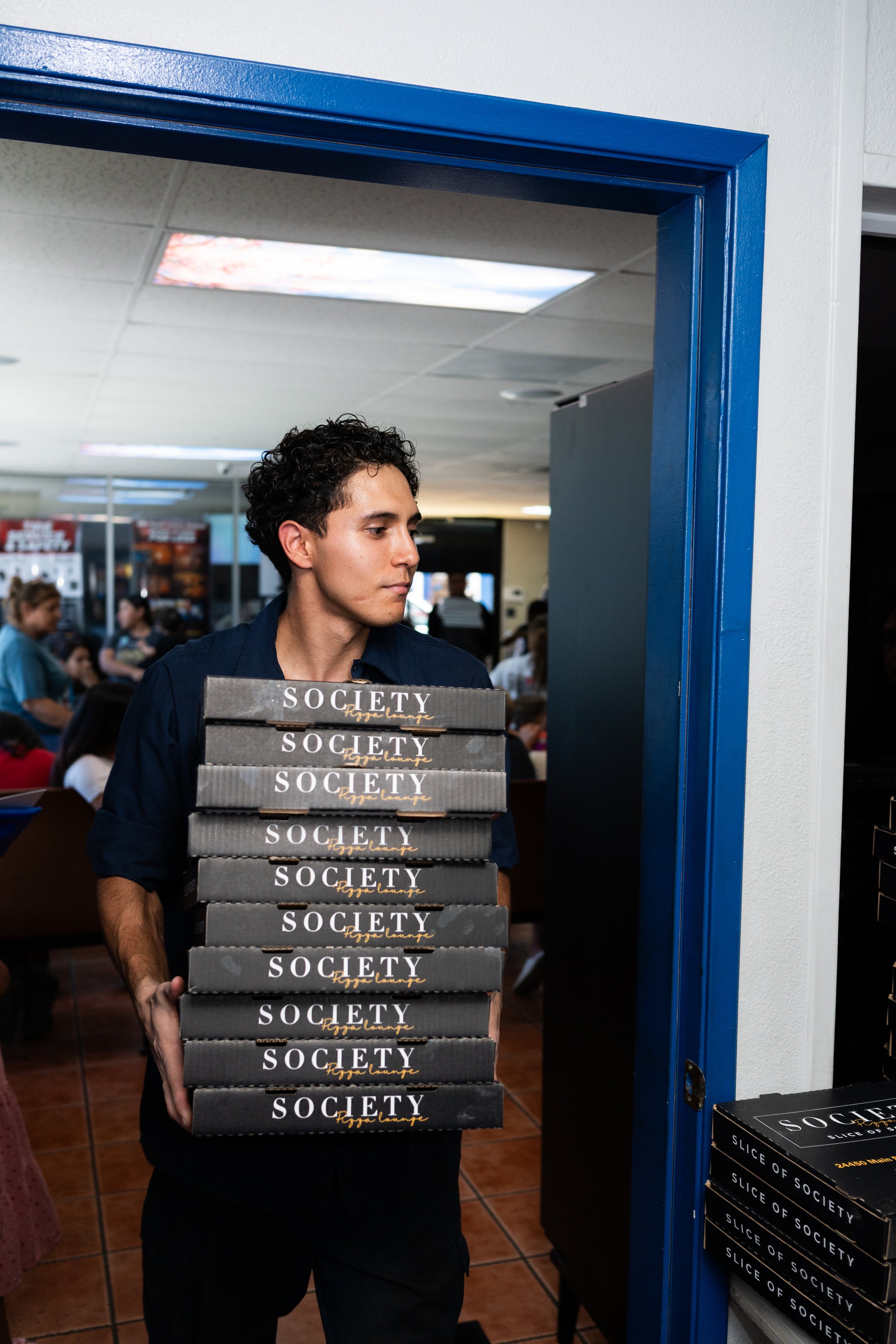A young man with dark, curly hair standing inside a restaurant or cafe, holding a stack of black boxes labeled 'SOCIETY' with yellow cursive writing, near a doorway painted blue.