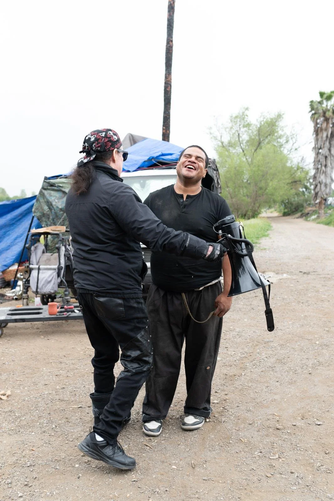 Two men are laughing and smiling, standing outdoors with camping gear and a tent in the background.