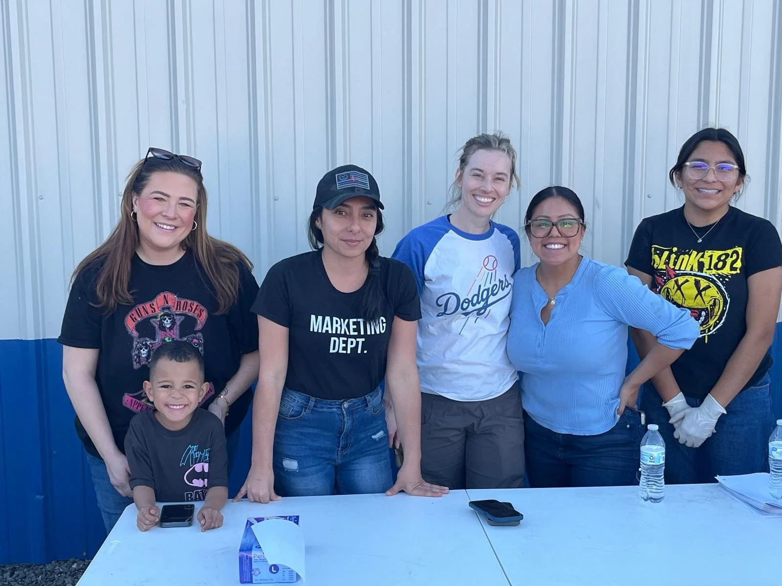 Group of six women and one child smiling and posing for a photo outdoors, standing behind a white table with water bottles and tissue box, with a corrugated metal wall in the background.