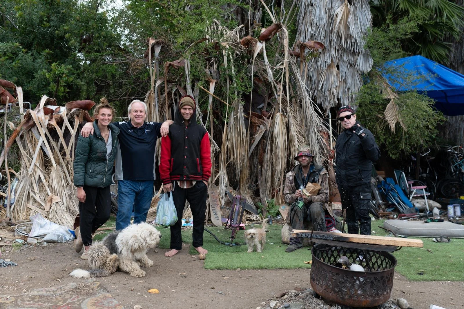 Group of five people and two dogs standing in front of a makeshift shelter made of dry palm leaves, with trees and bicycles in the background.