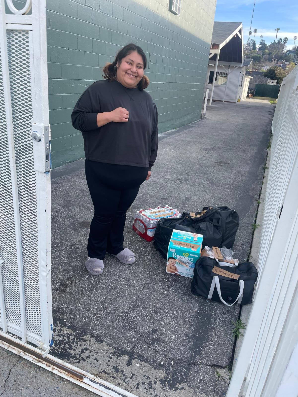 A woman standing outside next to a gate, smiling, with packages of diapers, food, and supplies on the ground beside her.