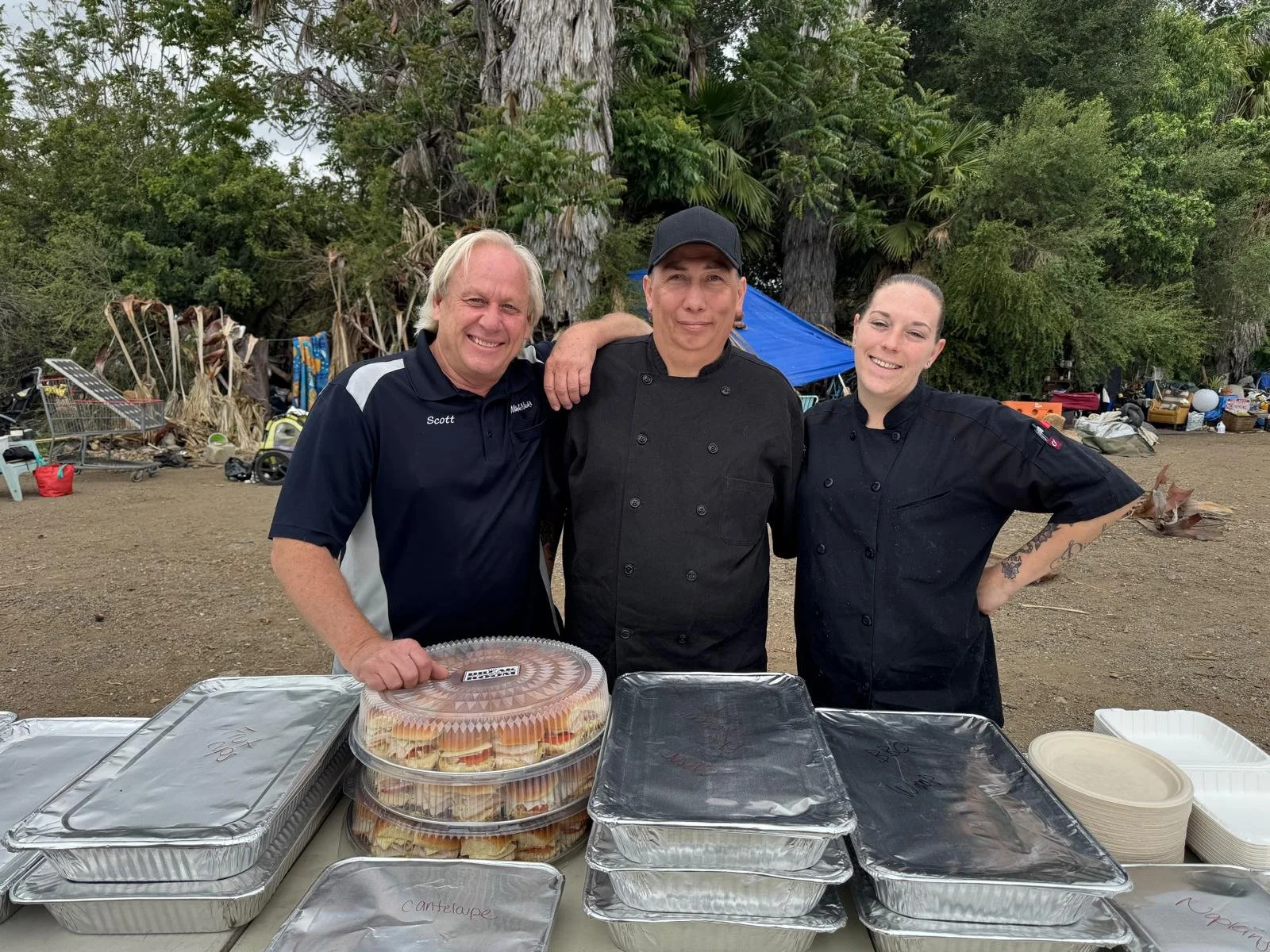 Three people, two men and one woman, standing behind a table with food containers, outdoors with trees and tents in the background.