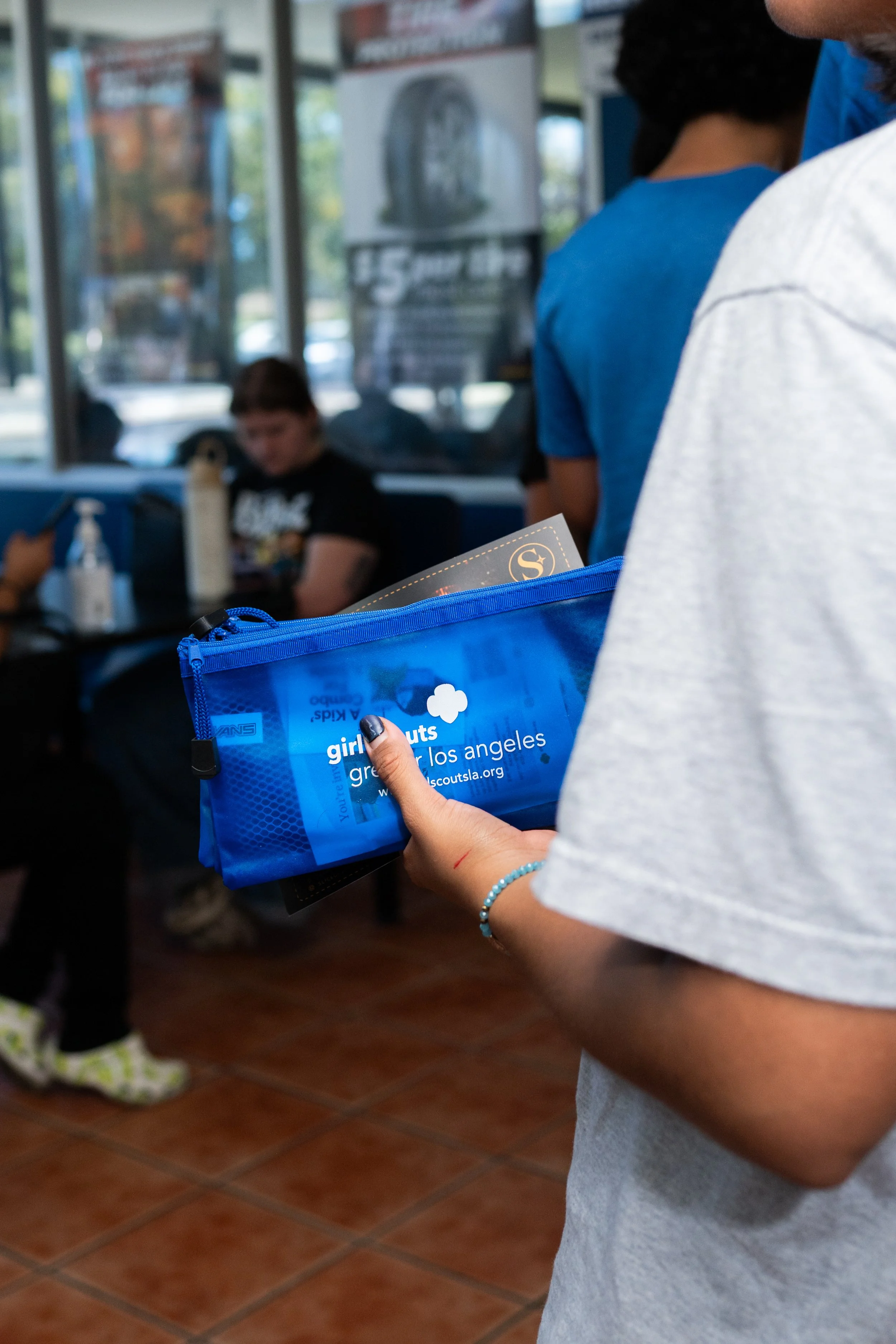 A person holding a blue bag with a Girl Scouts of America logo and a website, standing in line inside a store or community center. The background shows other people and posters.