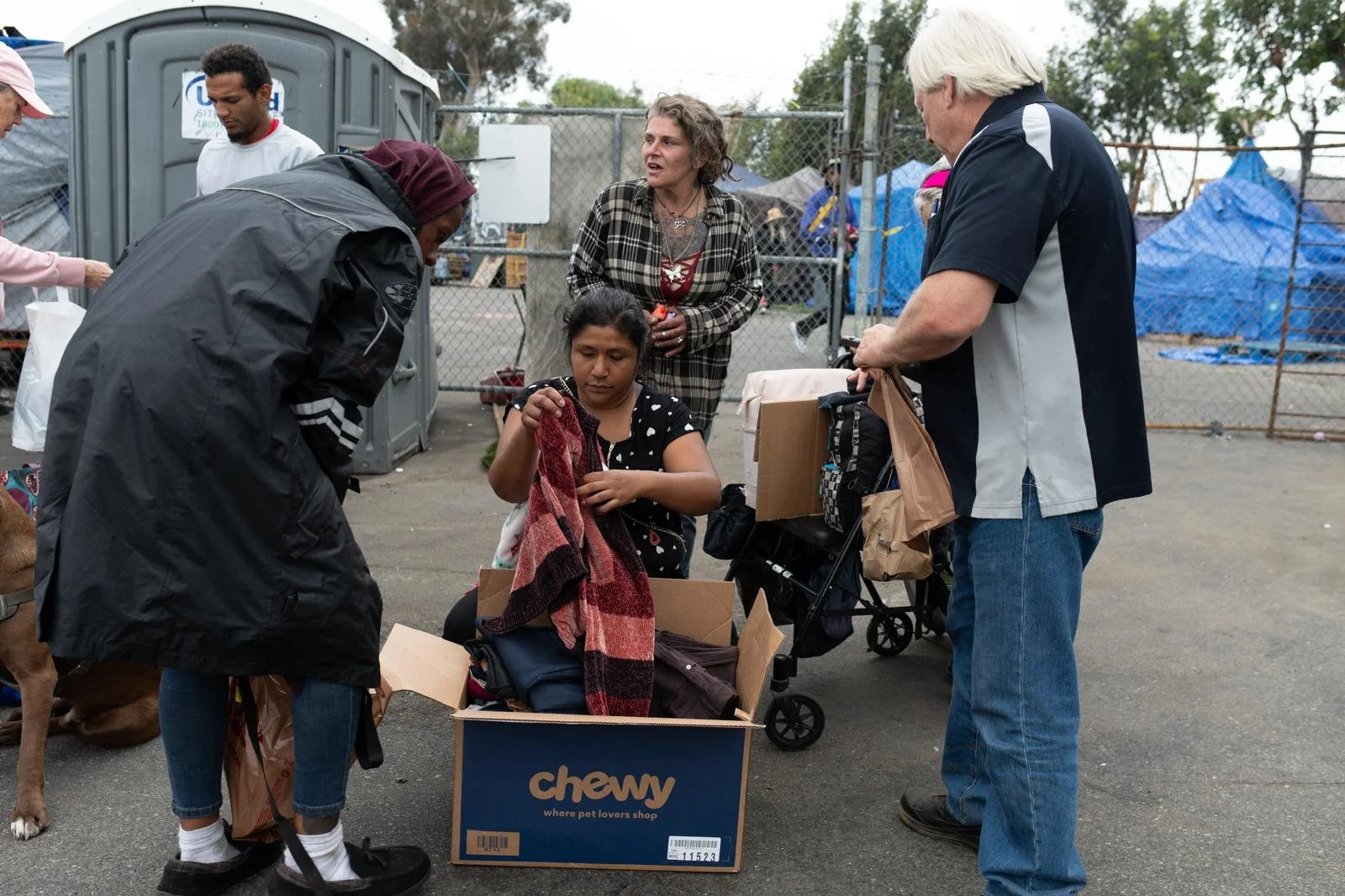 A group of people shopping at an outdoor market, some looking at clothing items and others engaging in conversation, with a fence and blue tarps in the background.