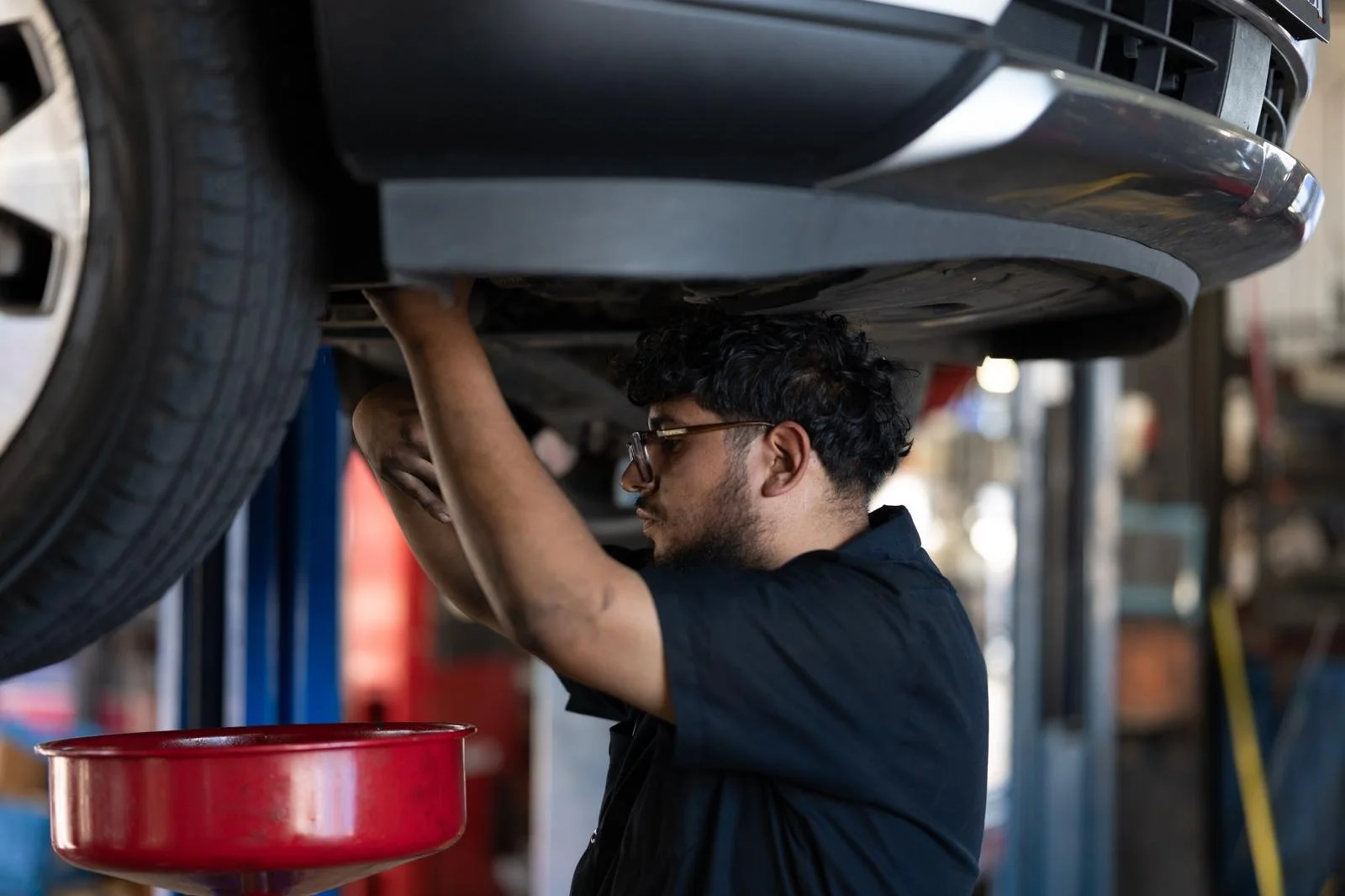 A mechanic working underneath a car on a lift, using his hands to perform maintenance. The mechanic is wearing glasses and a dark shirt, with a red container below him in a garage setting.