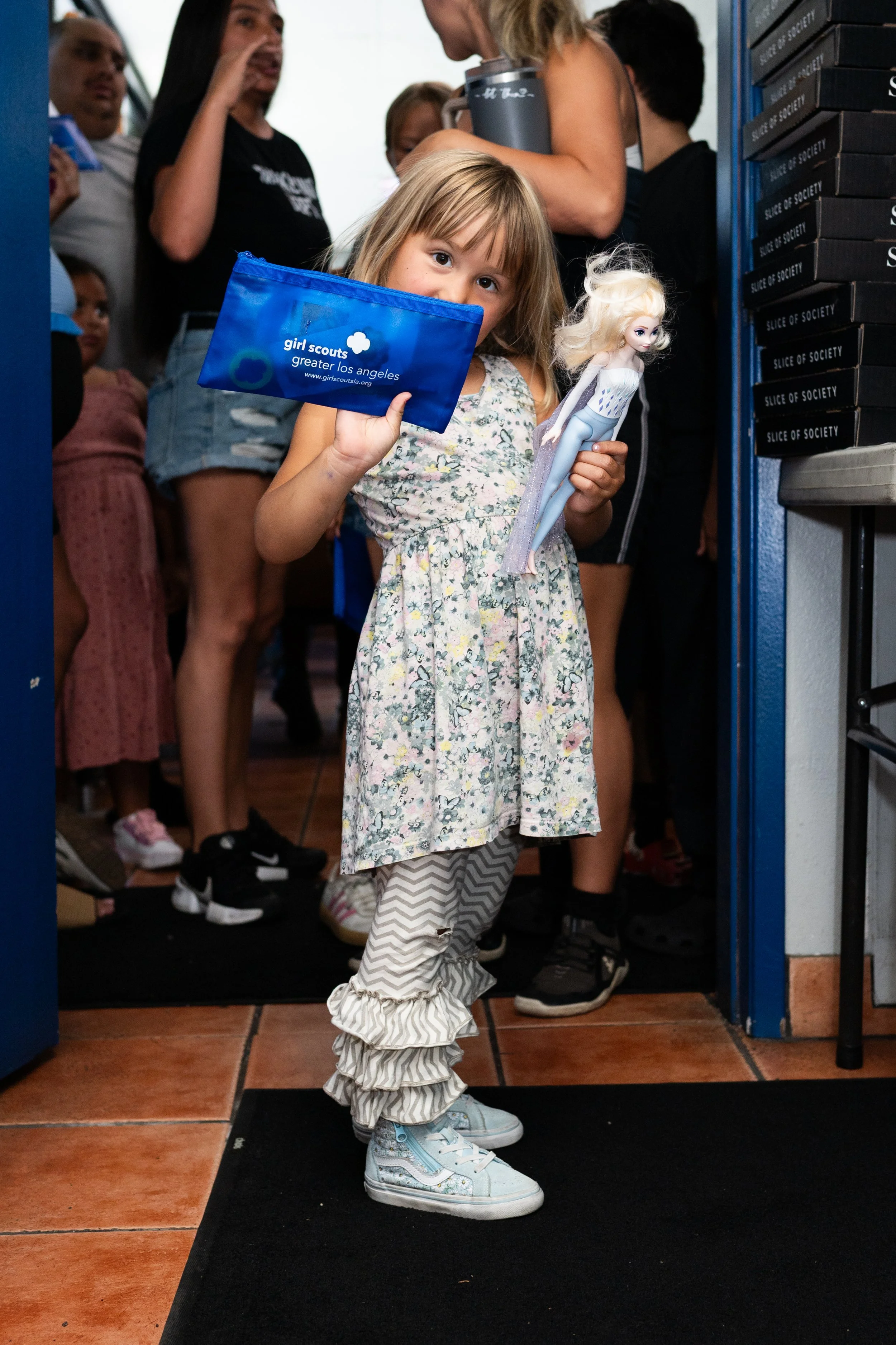 A young girl with blonde hair in a floral dress is standing on a mat inside a building, holding a doll and a Girl Scouts bag. There are several people behind her, some in line, some talking.