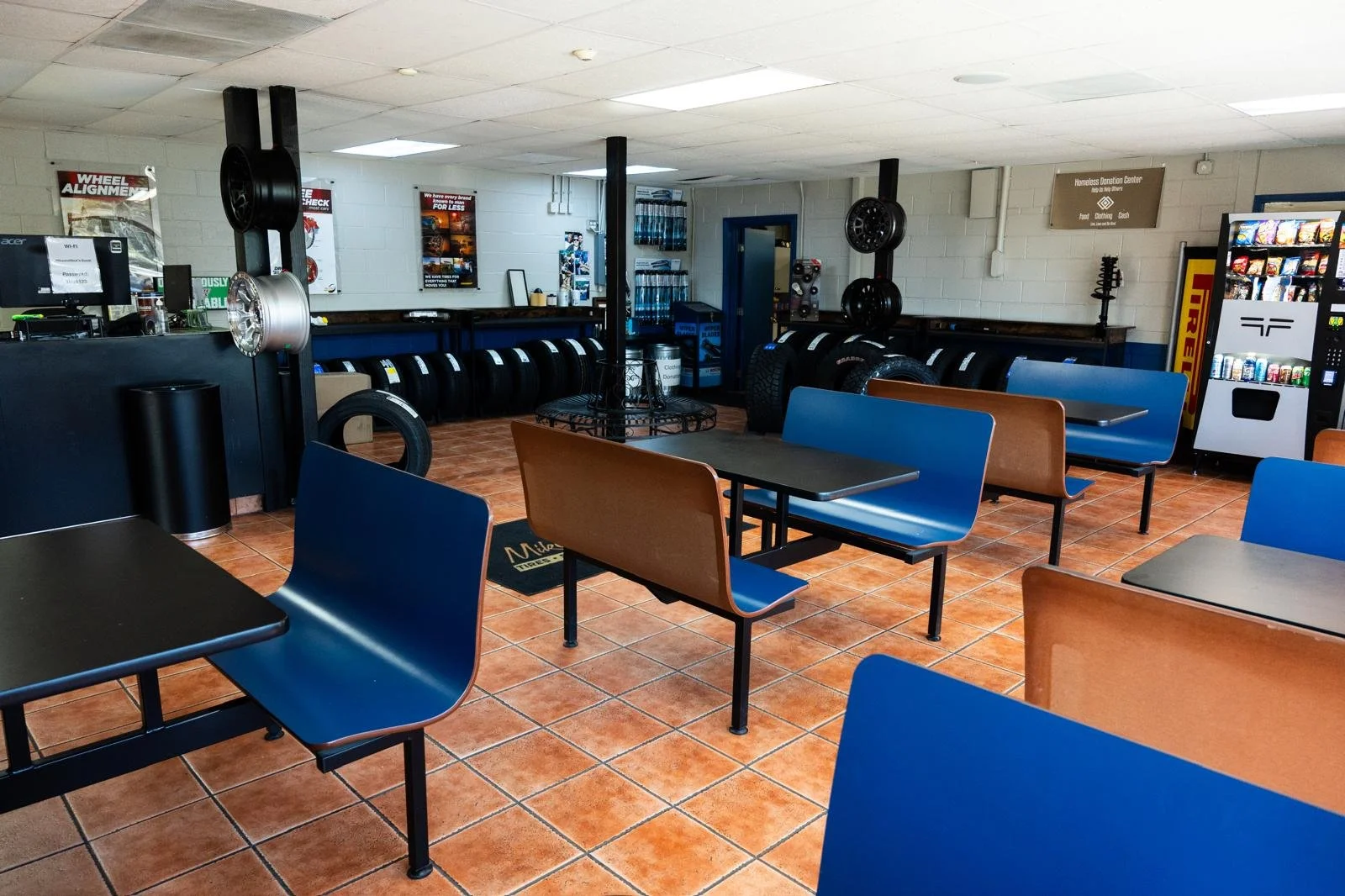 Interior of a waiting area with black tables and blue and brown chairs, tire racks, and a vending machine.