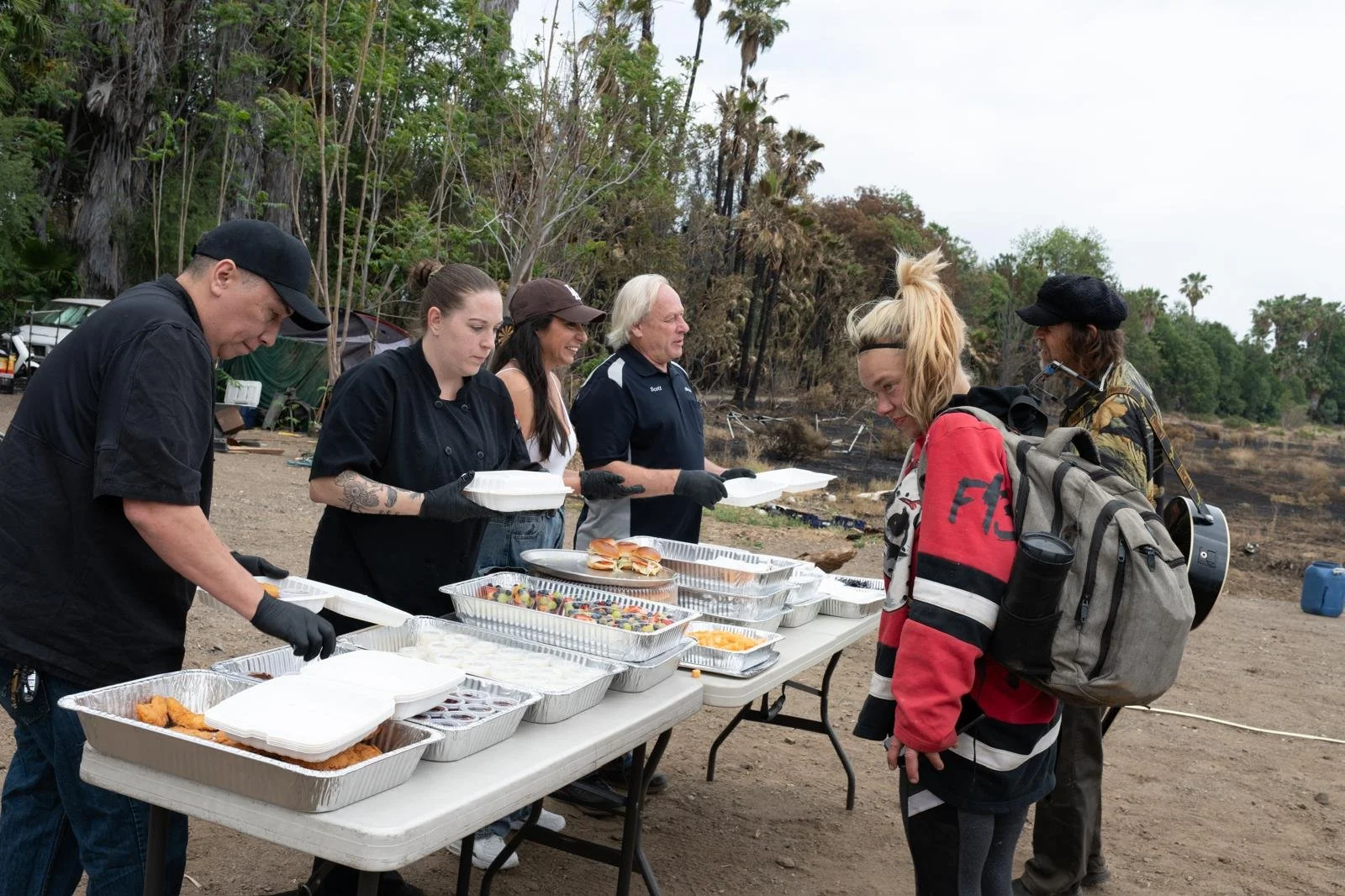 People serving and receiving food at an outdoor event with a dirt ground and trees in the background.