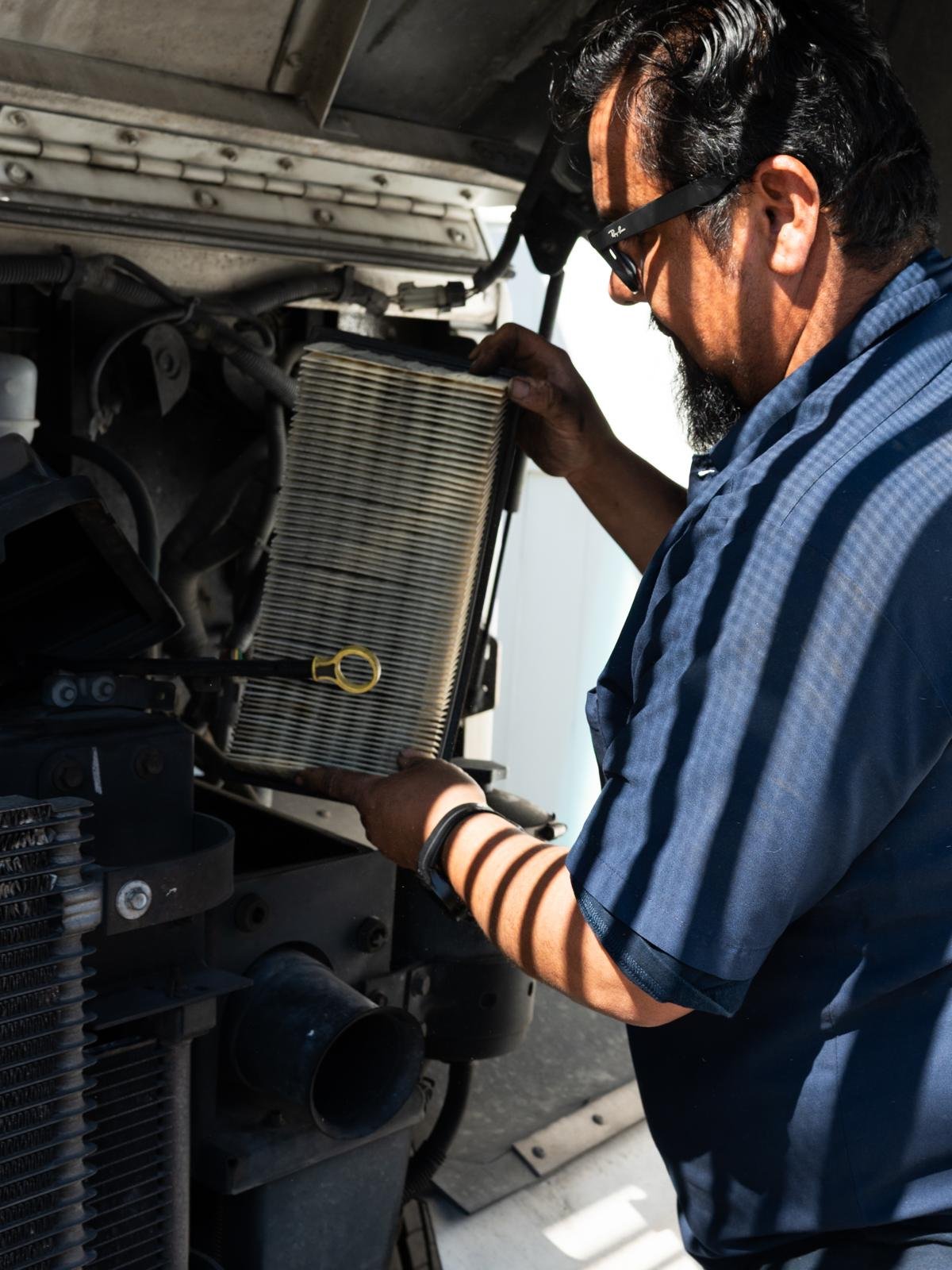 A man wearing sunglasses and a blue work shirt is working on a vehicle engine, holding a dirty air filter in his hands.