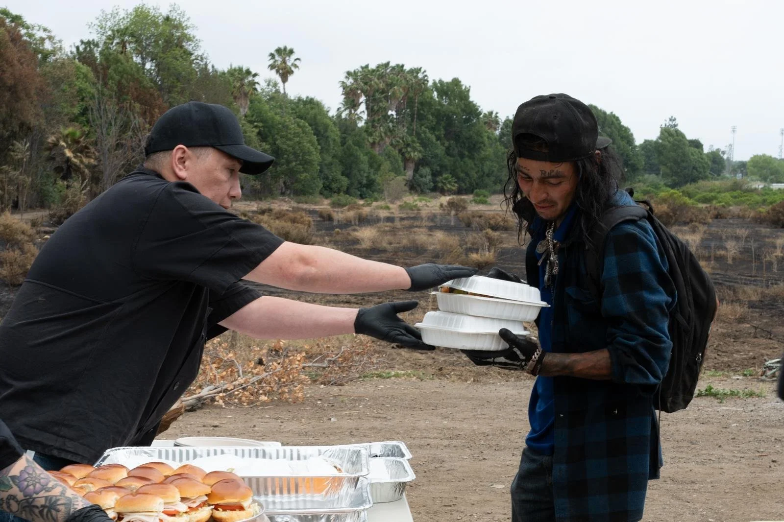 A person in black clothes and gloves is handing food containers to a man with a backpack, long hair, and a black cap, in an outdoor setting with trees and overcast sky.