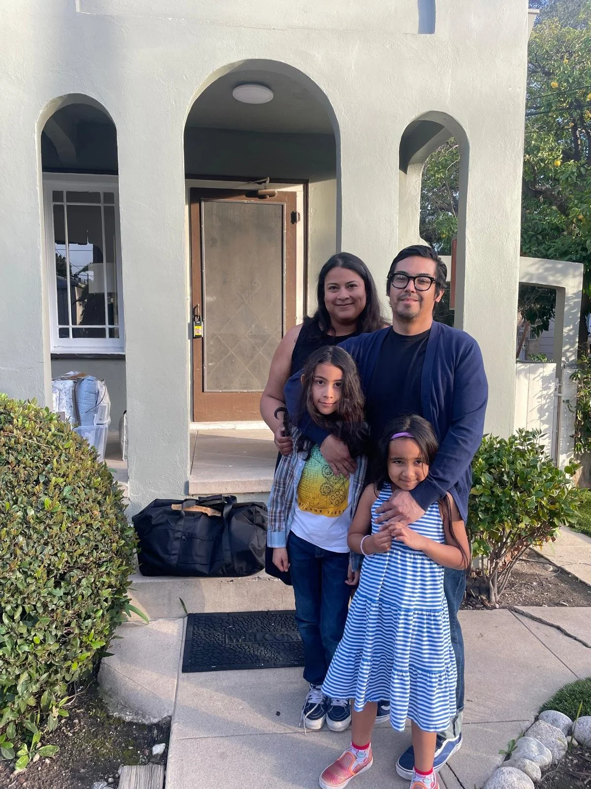 A family of four standing on a sidewalk in front of a house entrance, smiling at the camera. The house has an arched doorway and a light-colored exterior wall.