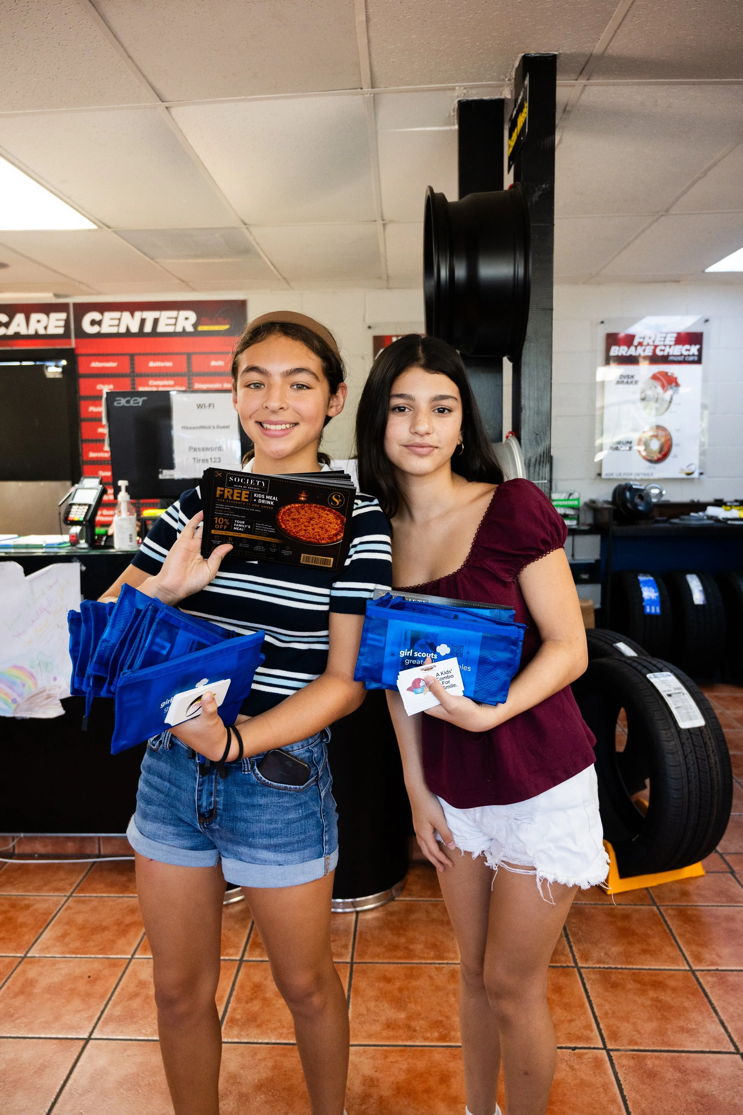 Two young girls standing inside a car service center, holding blue goody bags with Girl Scout logos. One girl is wearing a striped shirt and denim shorts, and the other is wearing a maroon top and white shorts. They are smiling and looking at the cam
