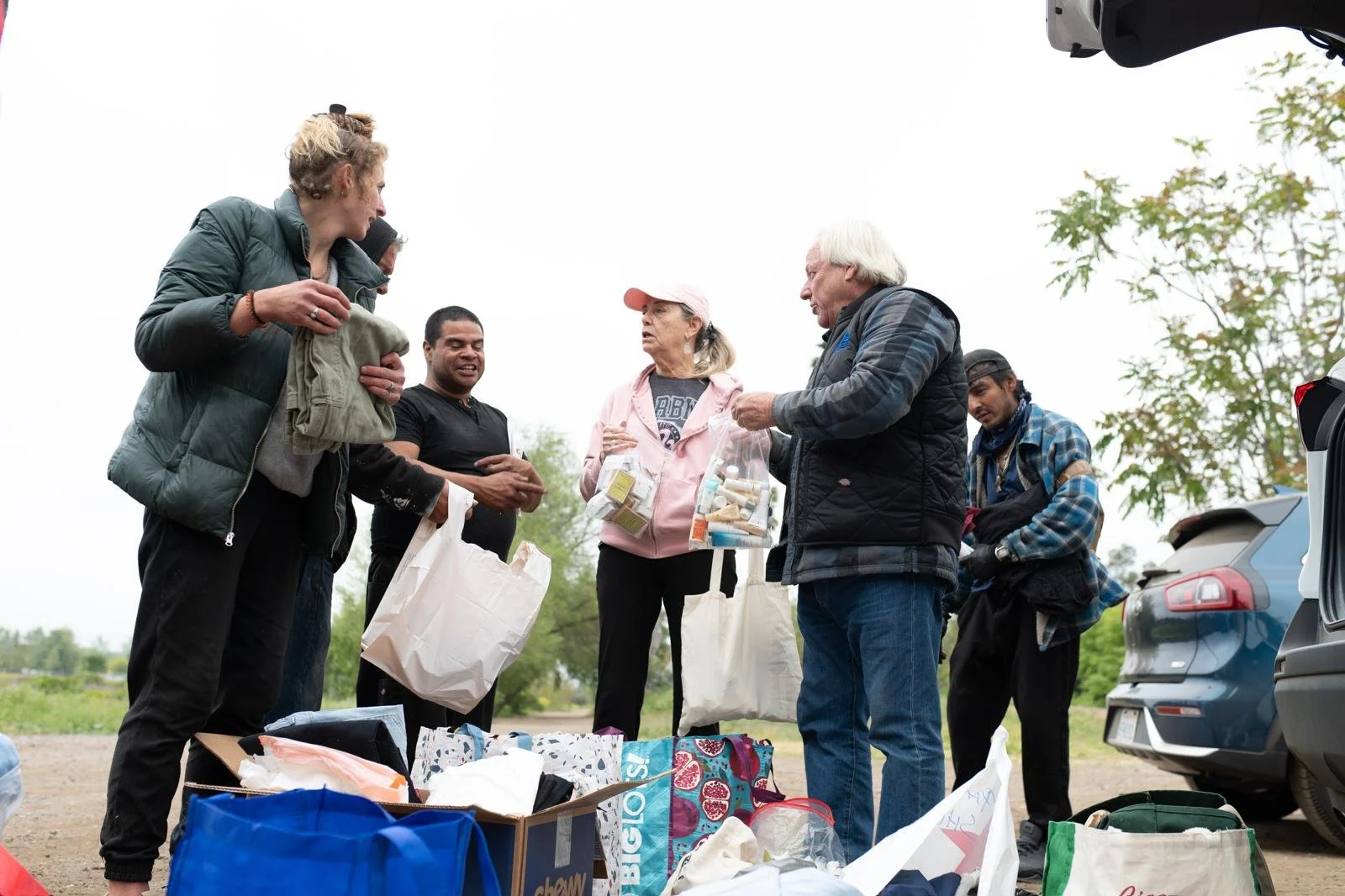 Group of people gathered outdoors around bags and boxes, some holding items and talking, in a casual setting with trees and cars in the background.