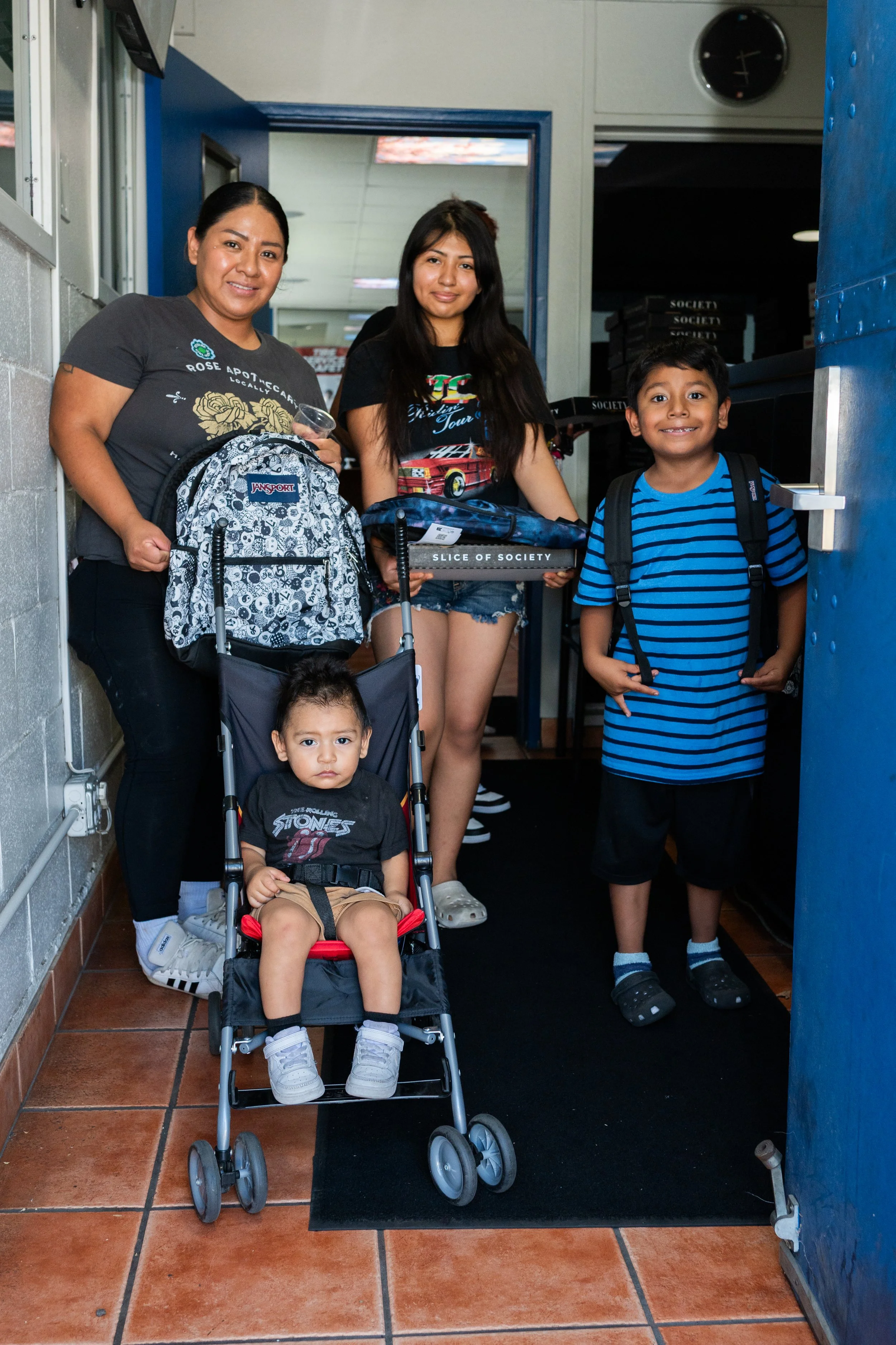 A group of people, including two women and three children, are standing and sitting in a doorway, smiling at the camera. The woman on the left is holding a backpack and a small container, the girl in the middle is holding a box, and the boy on the ri