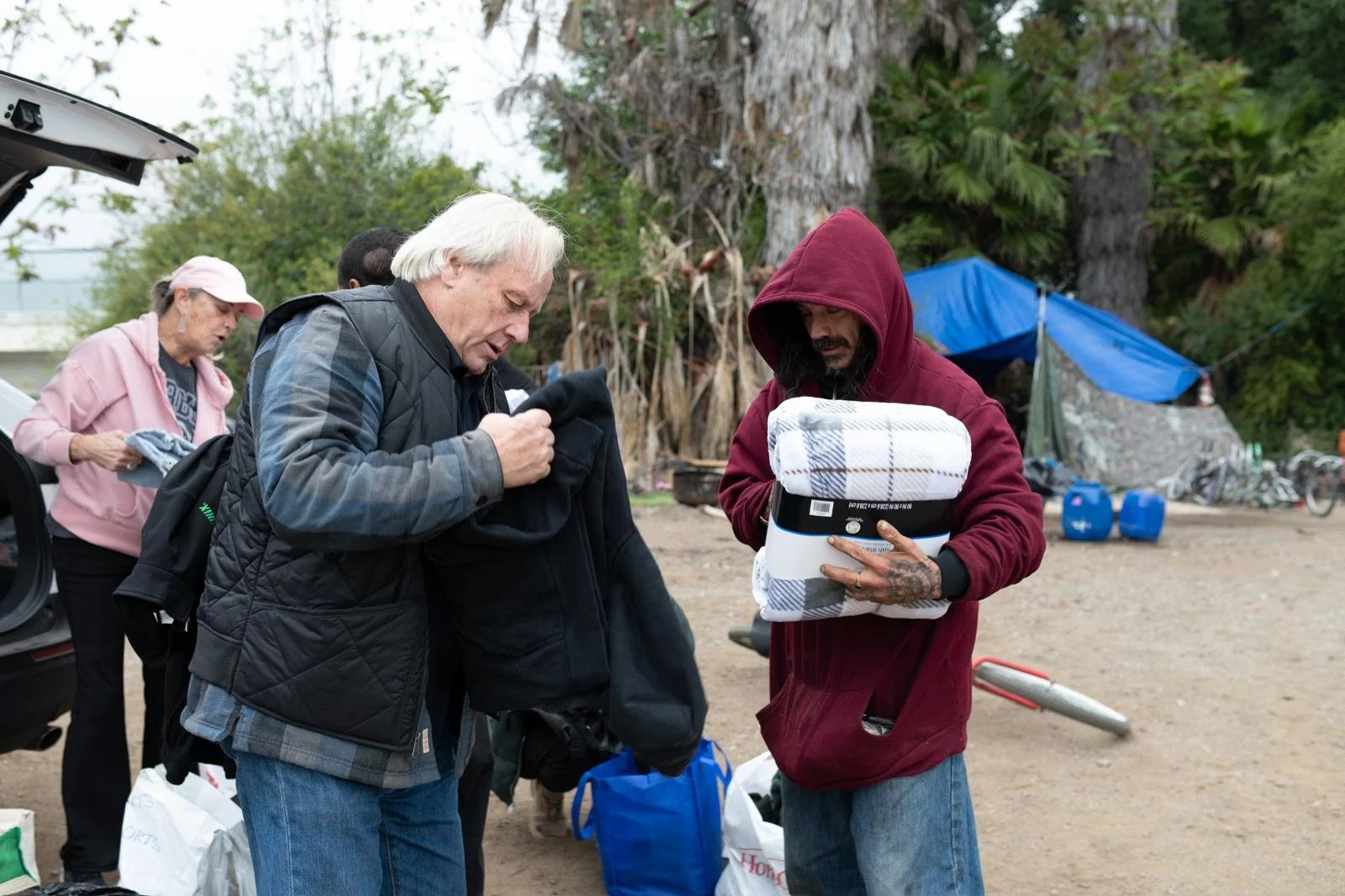A group of people at a campground, including a man with white hair and a black jacket, looking at an item. Another man with a red hoodie and tattoos, holding a folded blanket and a box, is standing nearby. A woman in a pink hat and pink jacket is in 