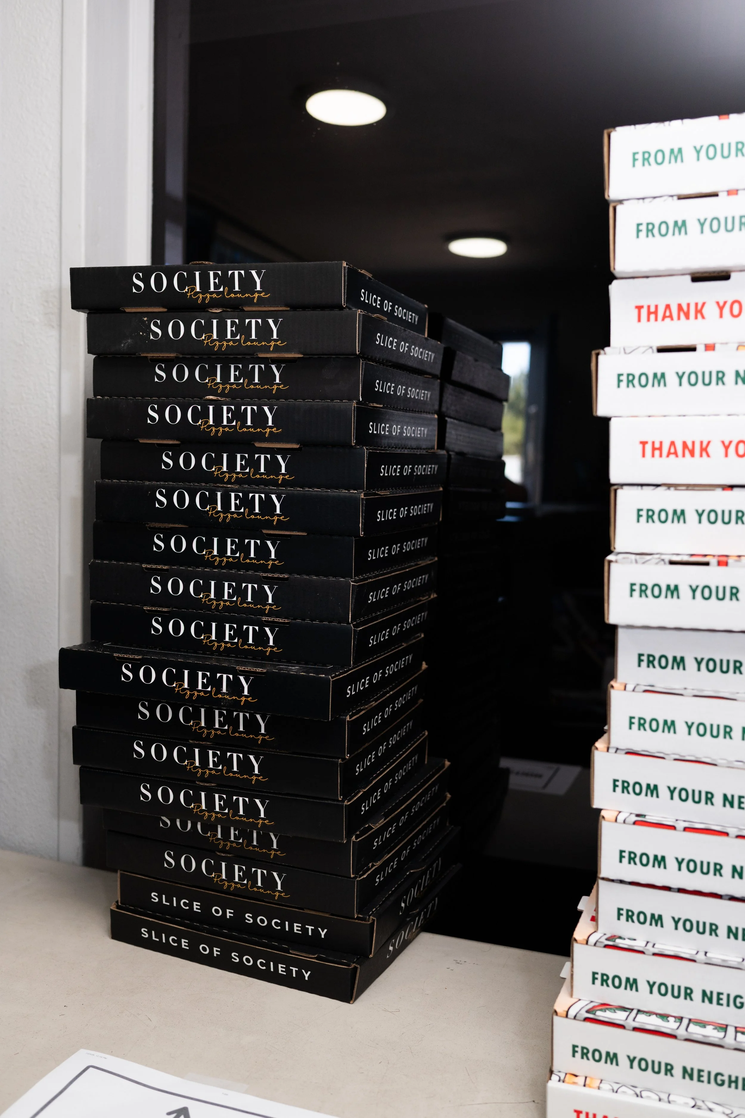 Stacks of black pizza boxes labeled 'SOCIETY Pizza Lounge' on a table and white boxes with 'FROM YOUR NEIGHBOR' and 'THANK YOU' printed on them in a store or distribution center.