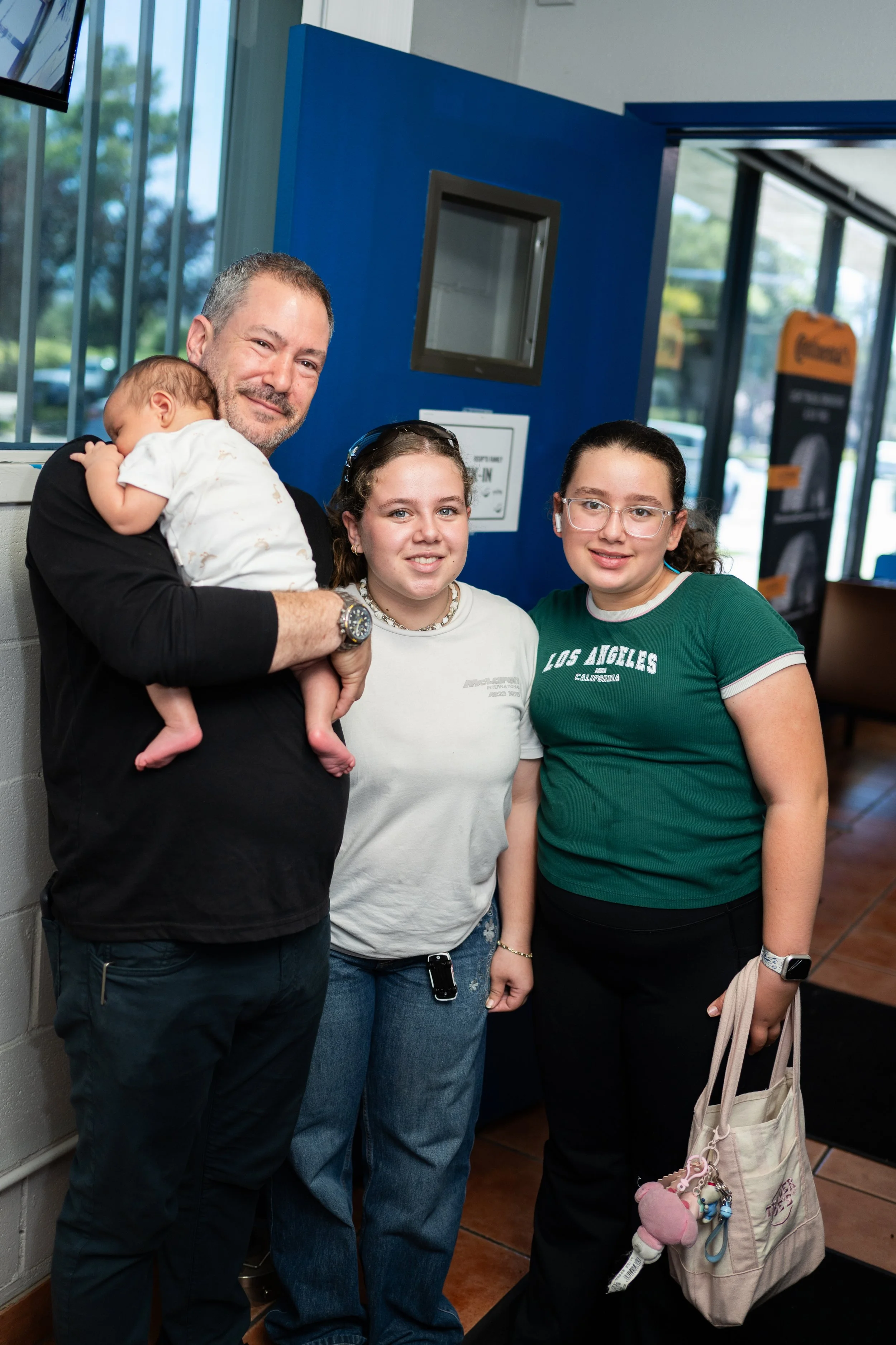 A group of four people, a man and three young women, pose together indoors near a blue wall and glass doors. The man, holding a baby, has short hair and a beard, and is smiling. The baby is sleeping on his shoulder. The young women have glasses and a
