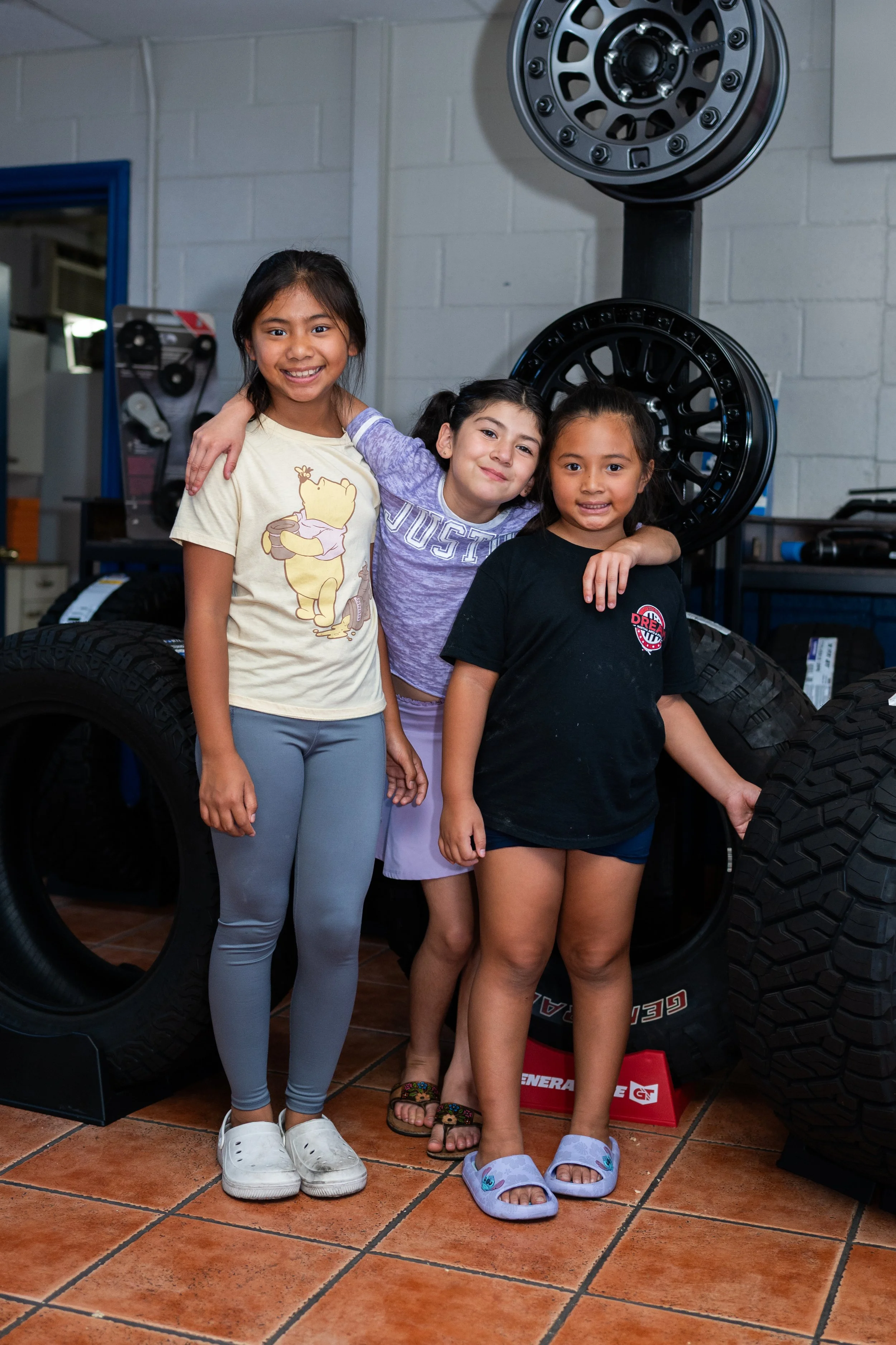 Three young girls standing together in a tire shop, smiling, with large tires and tools in the background.