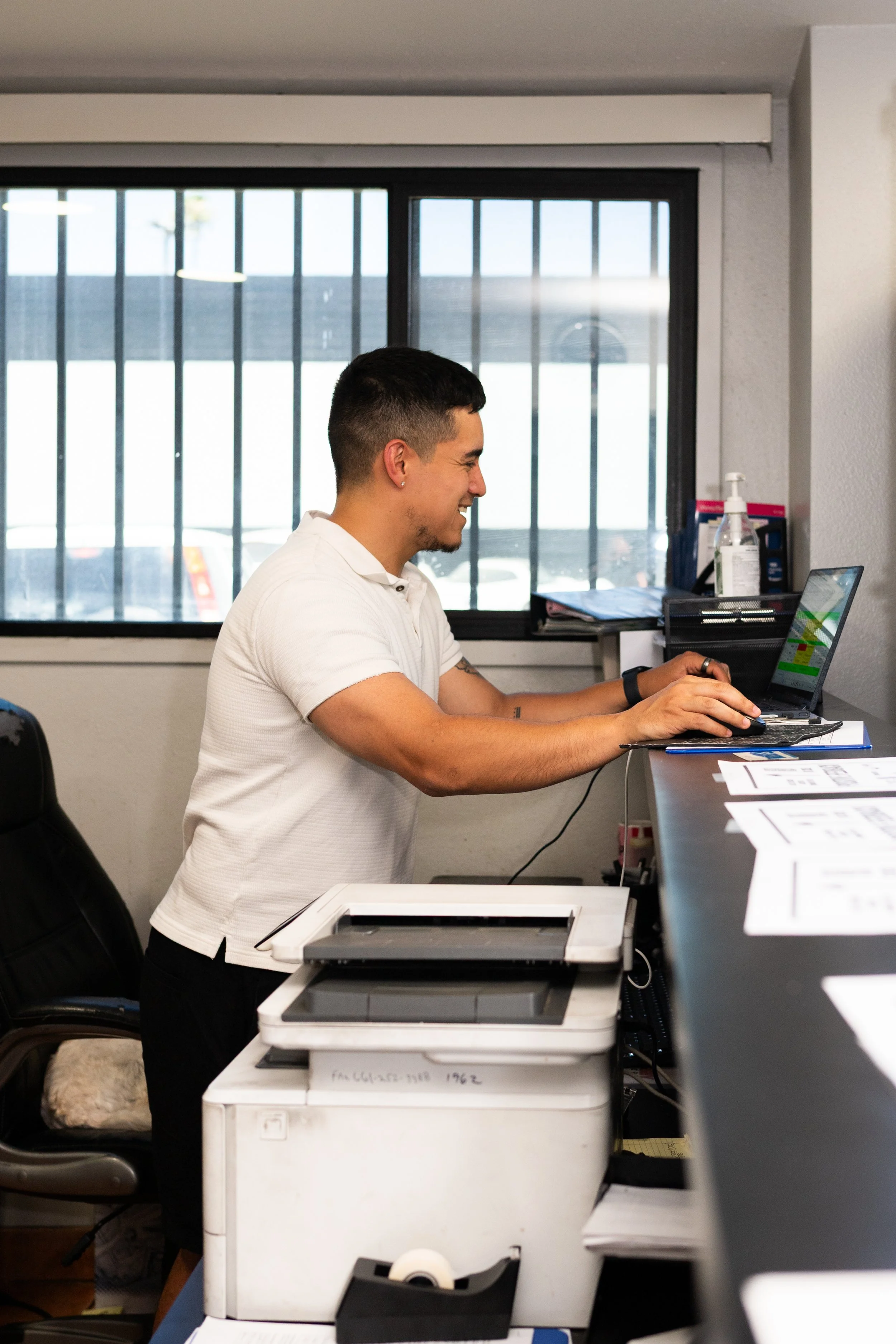 A young man with short dark hair, wearing a white polo shirt, is working on a laptop at a desk, smiling. The room has a large window with bars, and office supplies like papers, printers, and hand sanitizer are on the desk and nearby surfaces.