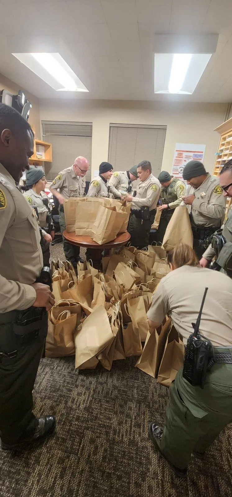 Group of uniformed officers in a room with brown paper bags on tables and floor, some officers are receiving or distributing items.