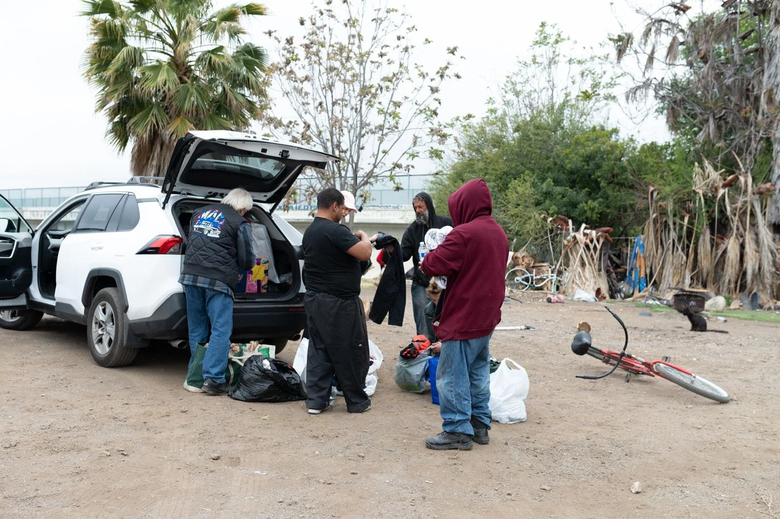 Group of people organizing supplies by a white SUV with open trunk on dirt ground, with trees and scattered debris in the background.