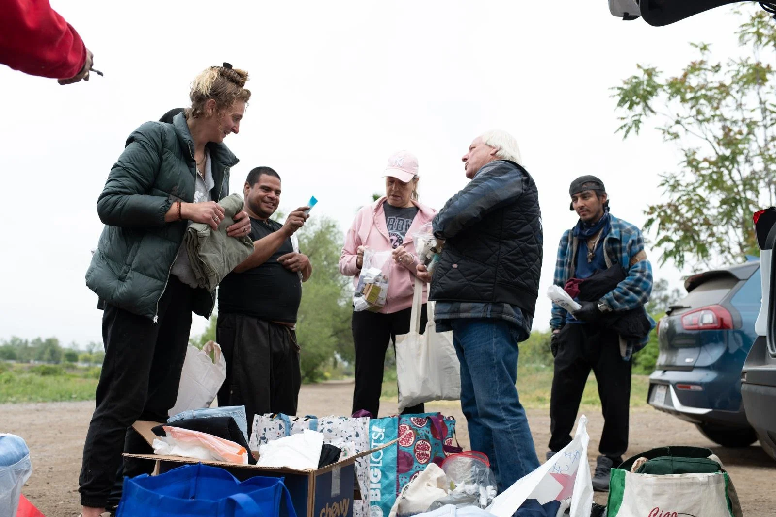 Group of people outdoors, standing around a pile of bags and boxes, engaged in conversation, with trees and cars in the background.