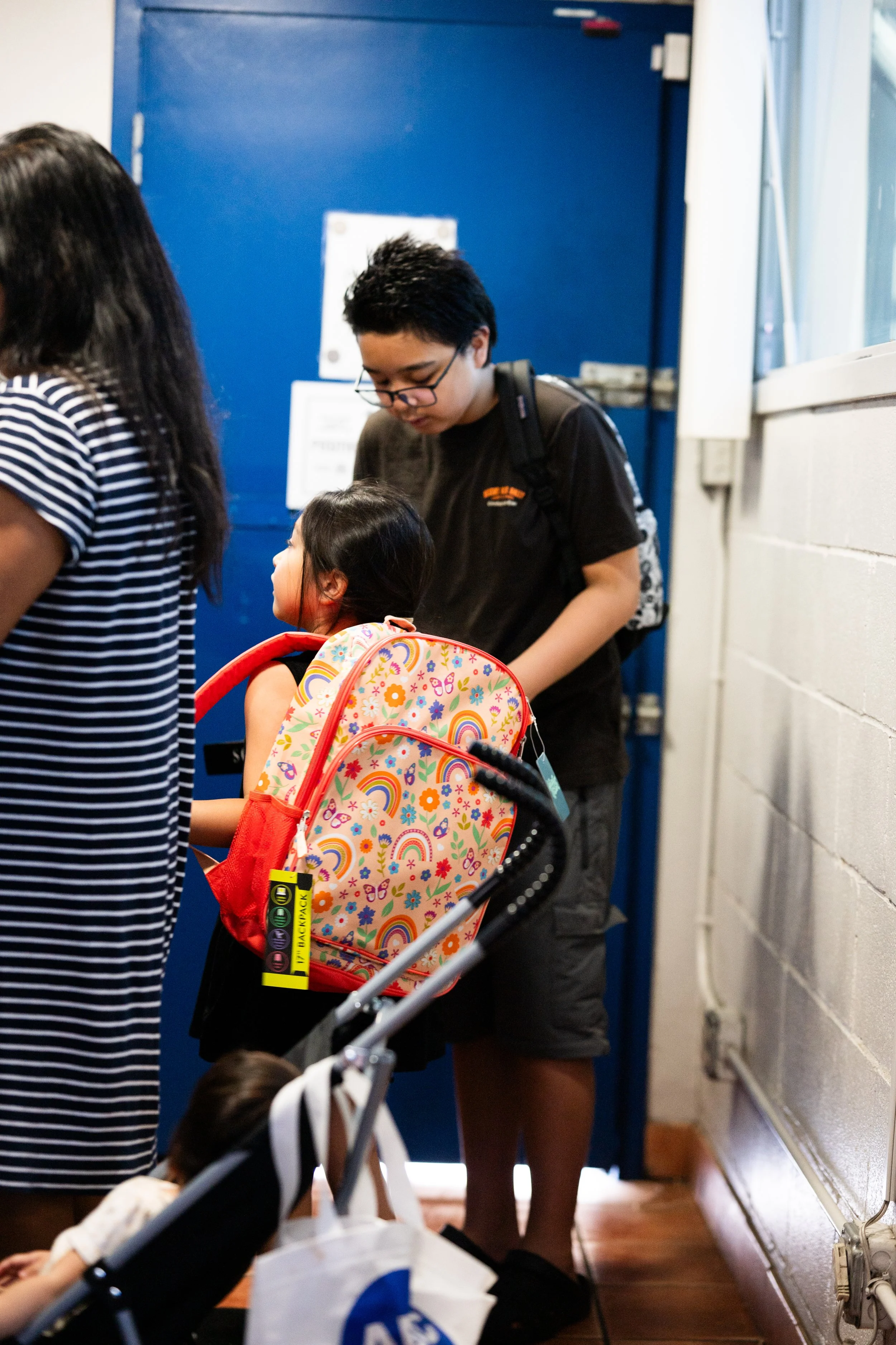 An adult with short black hair and glasses is interacting with a young girl with long black hair, who is wearing a colorful rainbow-themed backpack. The scene appears to be in a hallway near a blue door. Other individuals are present but mostly out o