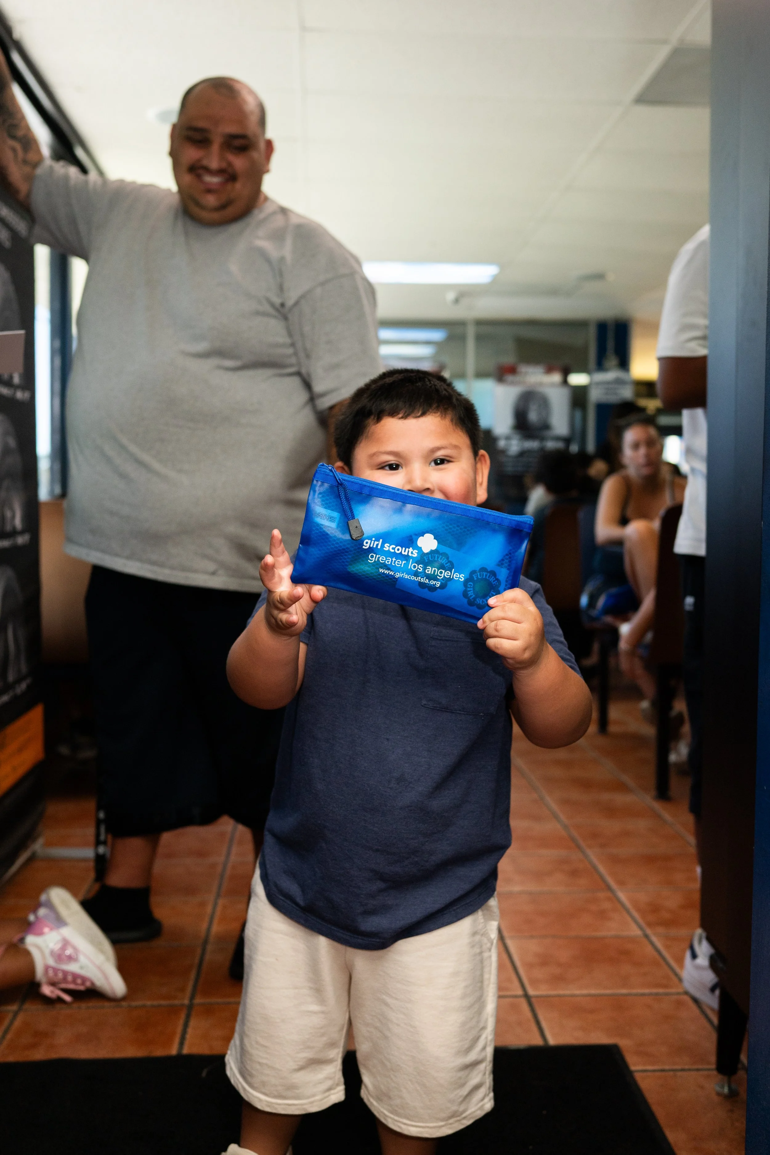 Young boy holding a Girl Scouts of Greater Los Angeles blue pouch, smiling, indoor restaurant setting.