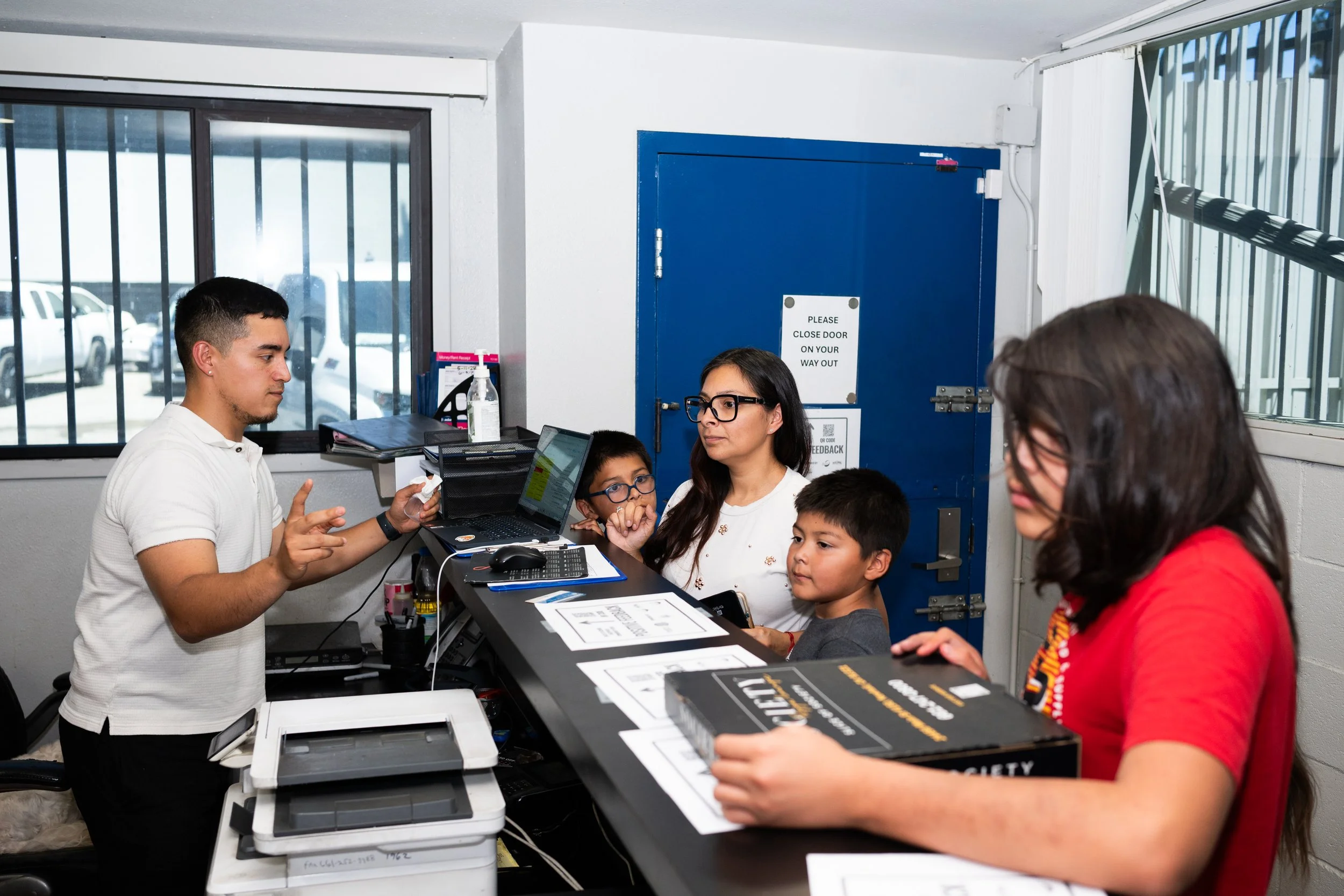 A group of people inside a room near a counter, with a woman, a man, and two children visible. The man appears to be explaining something, with a laptop and a printer on the counter in front of him. The wall behind has a blue door with a sign that sa