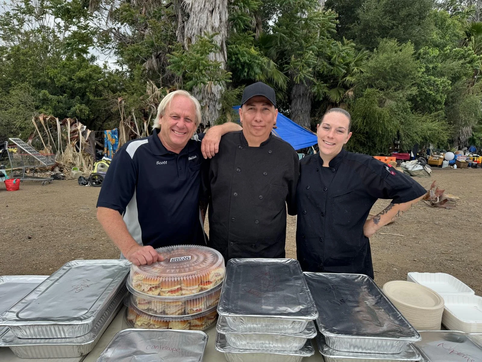 Three people, two women and one man, stand behind a table filled with food containers outdoors, with trees and tents in the background.