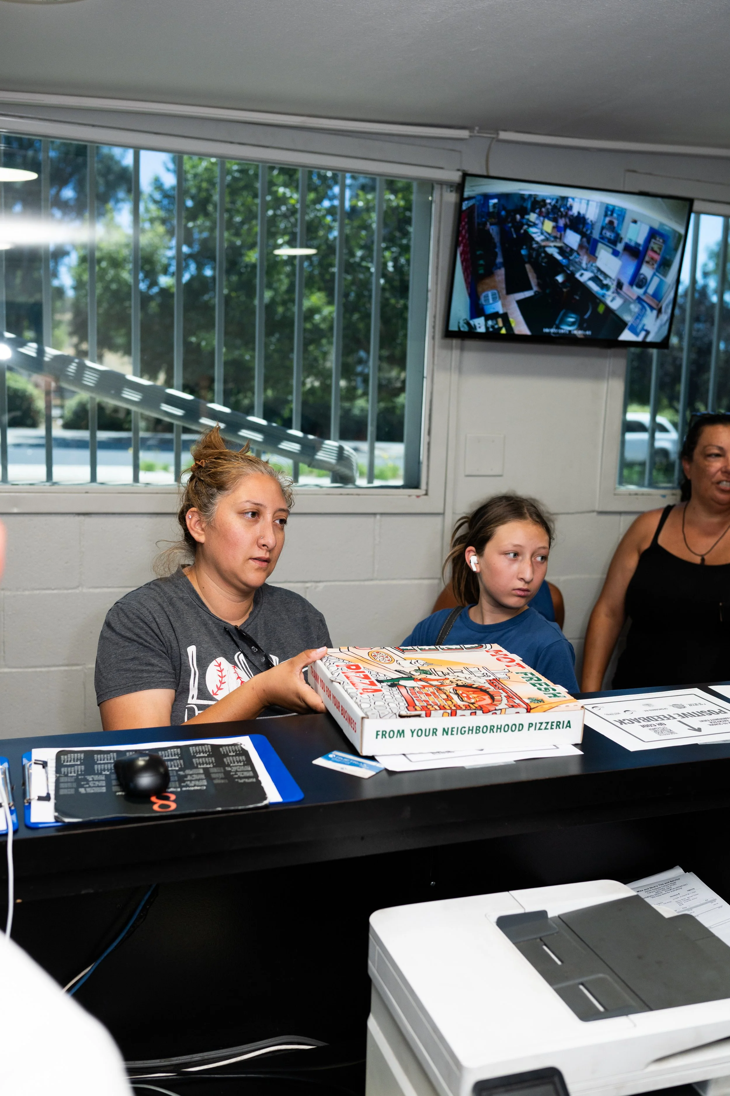 People sitting at a table in a room with large windows, with a woman holding a pizza box, a computer mouse, a keyboard, and a printer on the table.