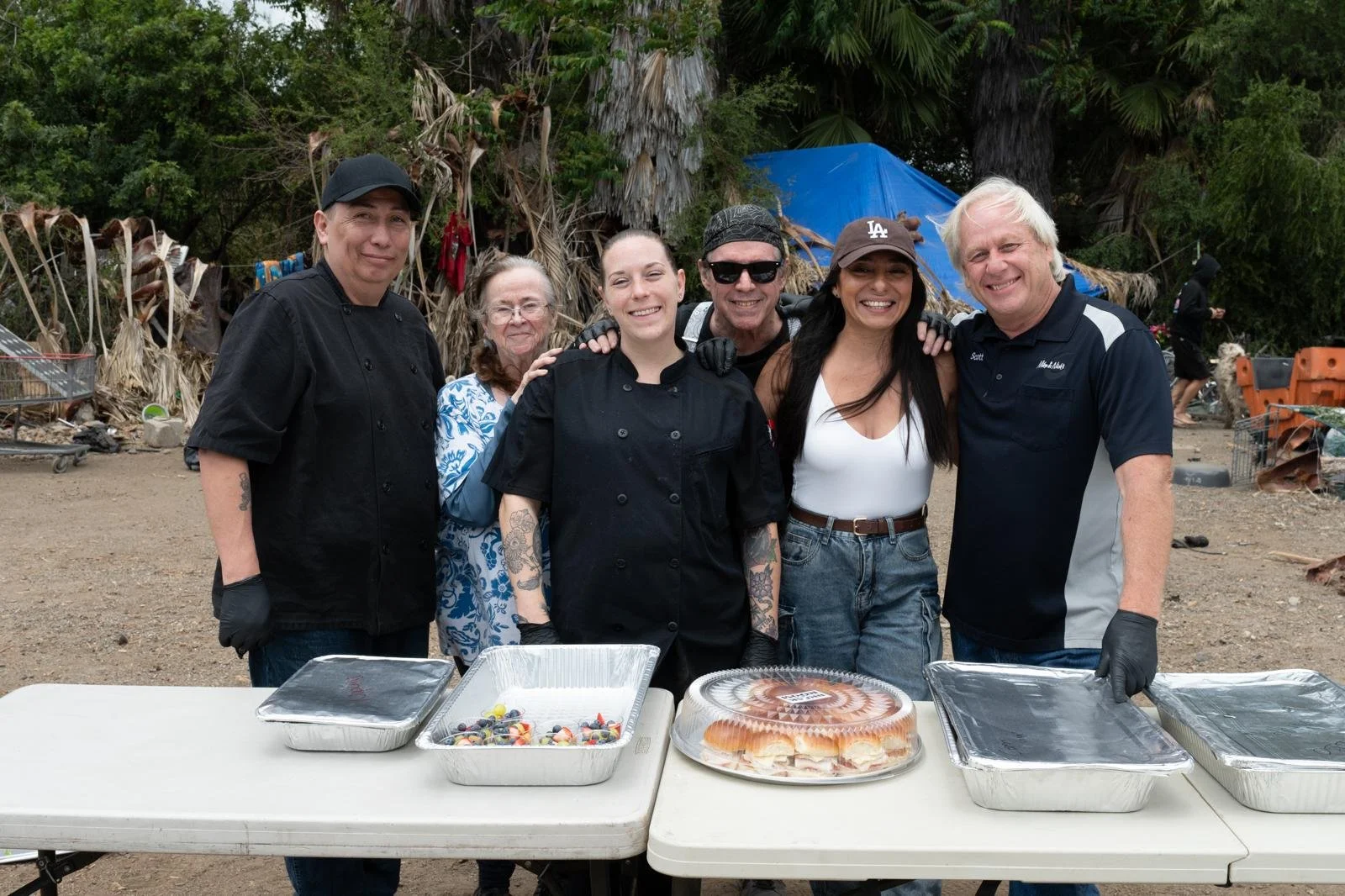 Group of six people standing outdoors behind a table with food, smiling, with trees and a makeshift shelter in the background.
