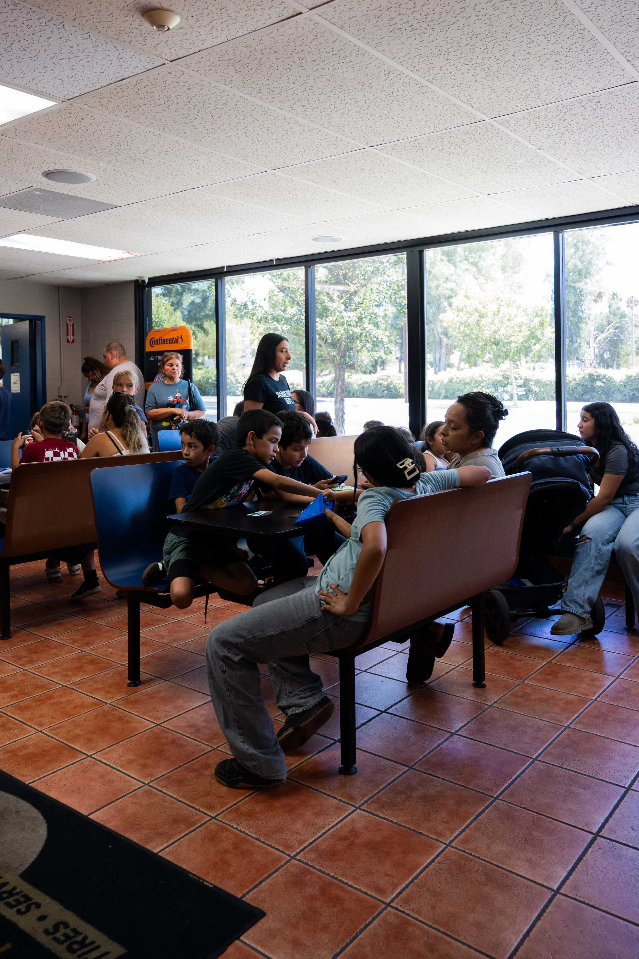 A group of children sitting on chairs in a waiting area, while several adults stand around, with large windows showing trees outside.