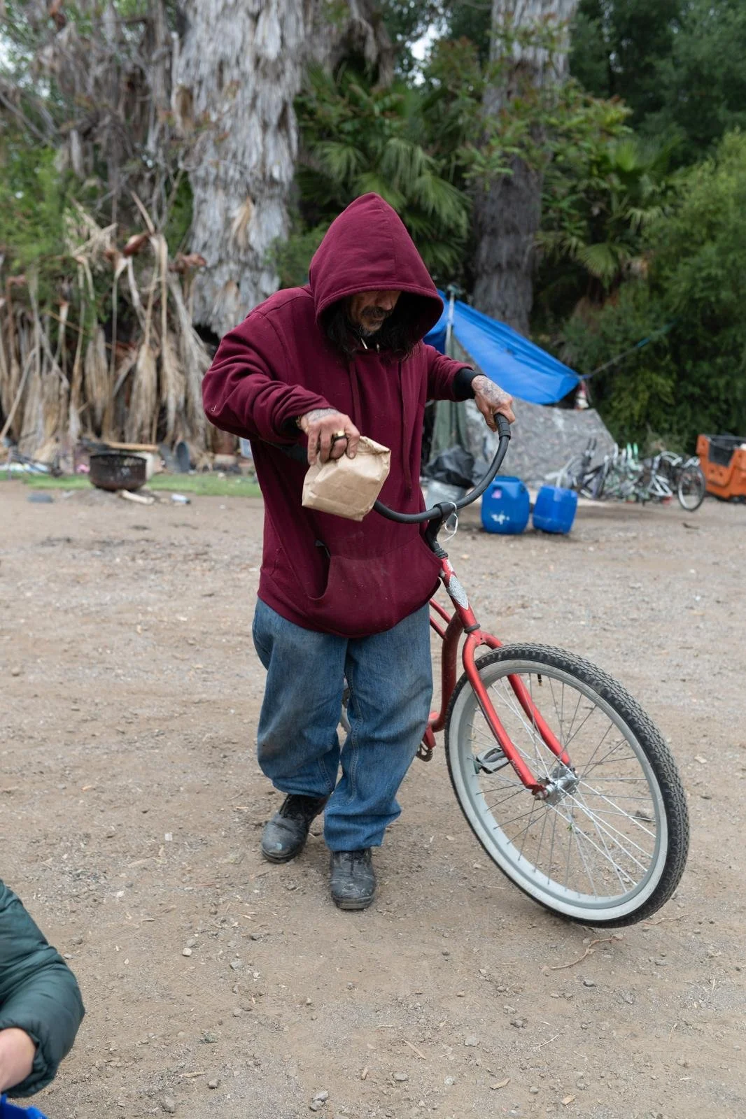 A man in a red hoodie and baggy jeans fixing a red bicycle on a dirt lot surrounded by trees, with some blue barrels and a tarp in the background.