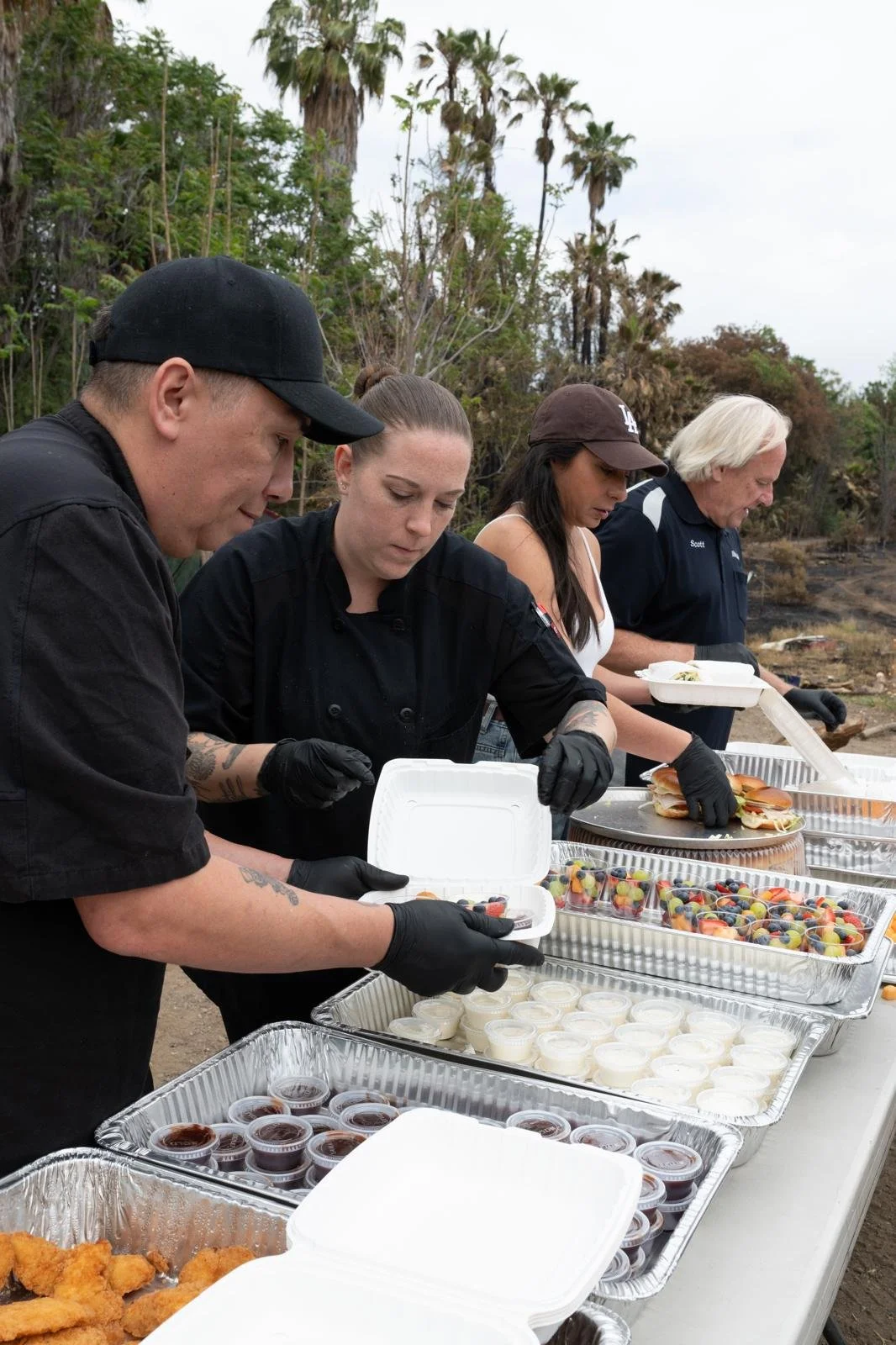 People serving food at an outdoor table with various desserts and condiments.