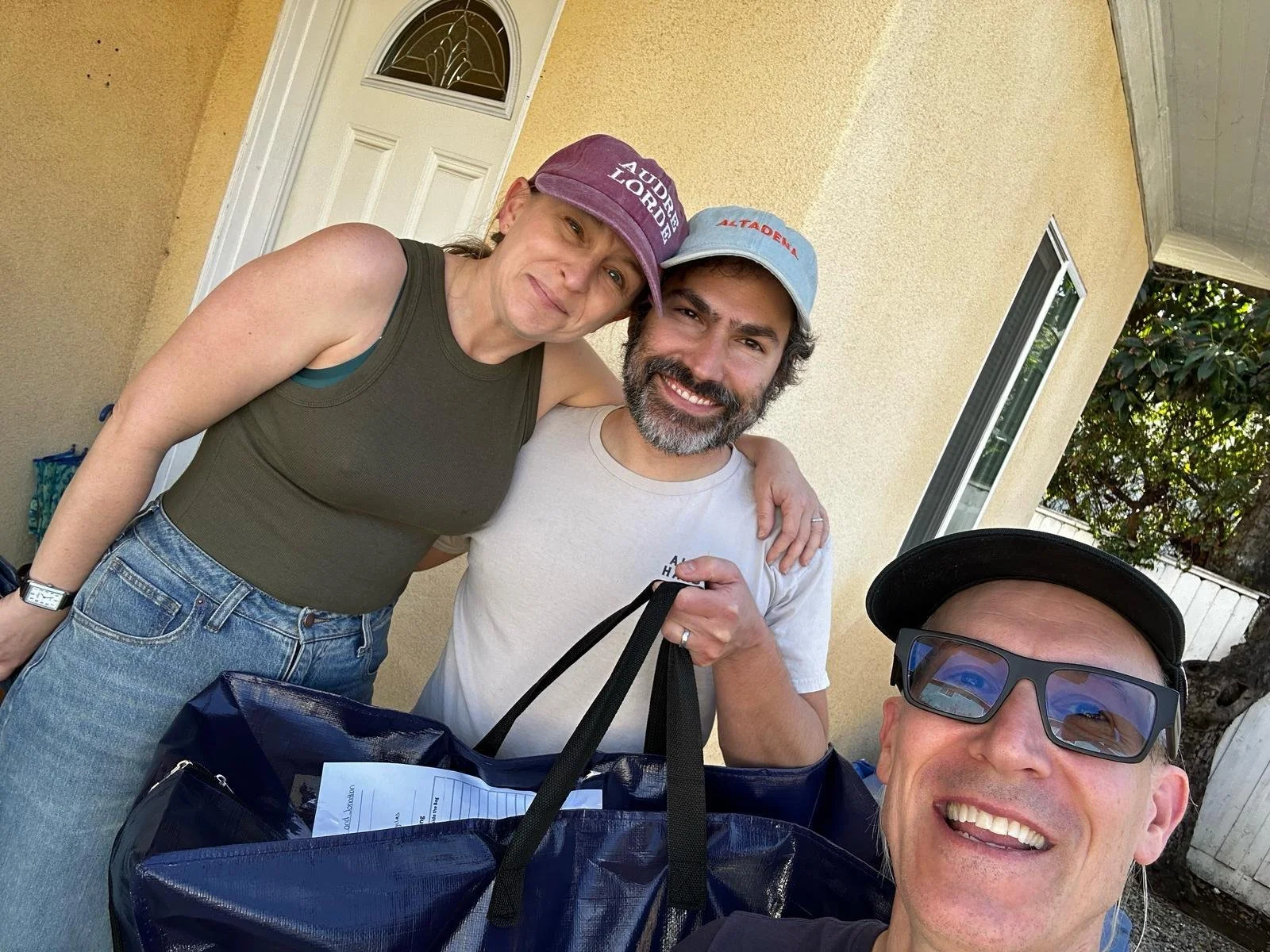 Three people smiling and posing for a selfie outside a house, with one person holding a bag and papers.