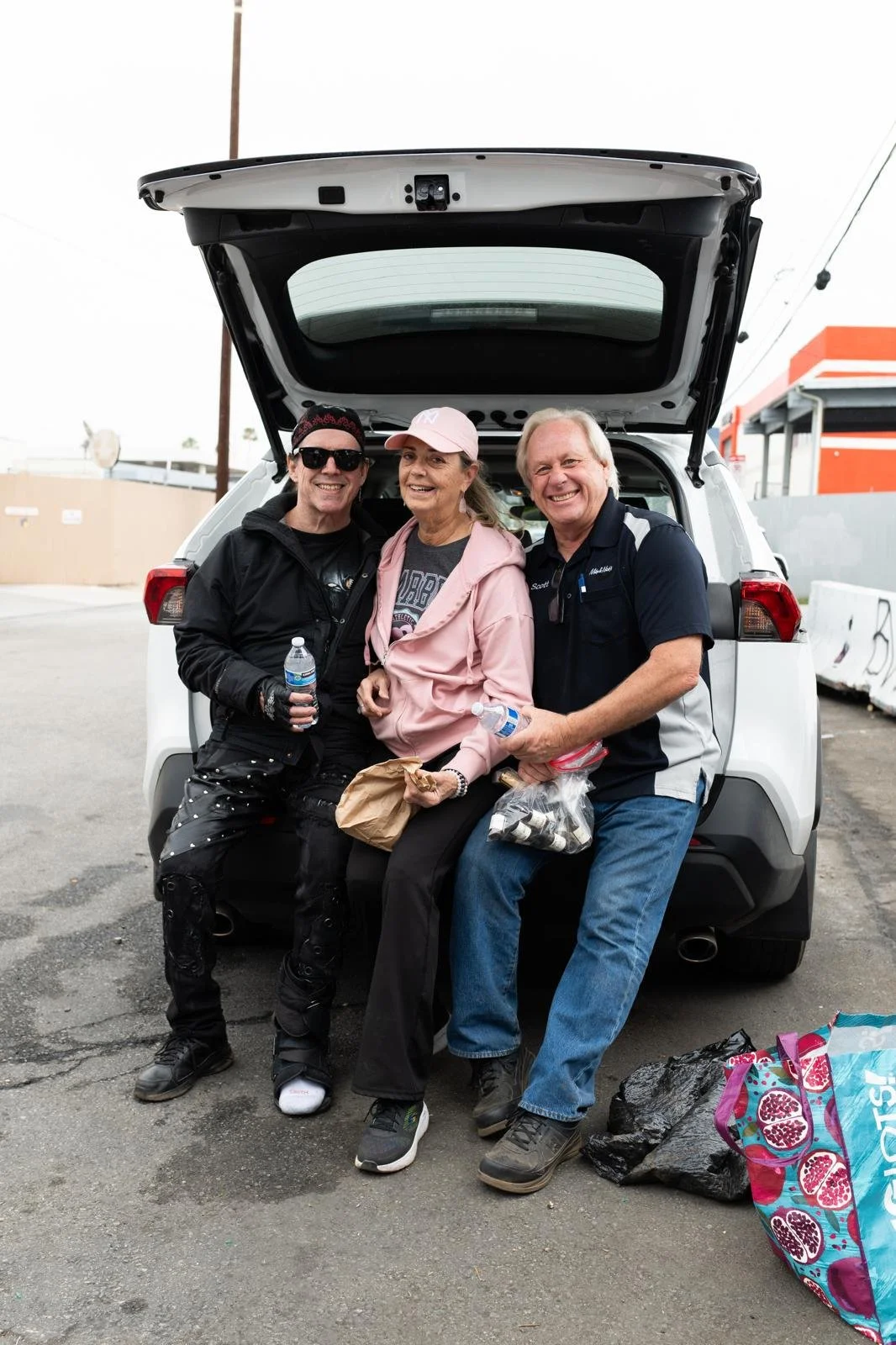 Three people sitting on the open back of a white SUV, smiling, in an outdoor parking lot. The person on the left is wearing black clothing and sunglasses, holding a water bottle. The person in the middle is wearing a pink jacket and cap, holding a br