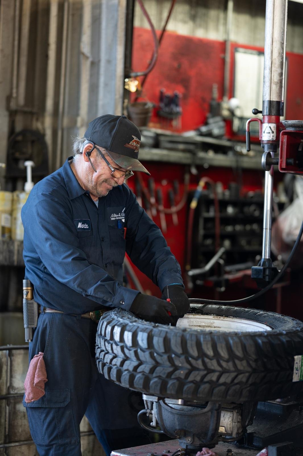 A mechanic working on a tire in a garage or repair shop, wearing a navy blue uniform, glasses, and a black cap, with various tools and equipment in the background.