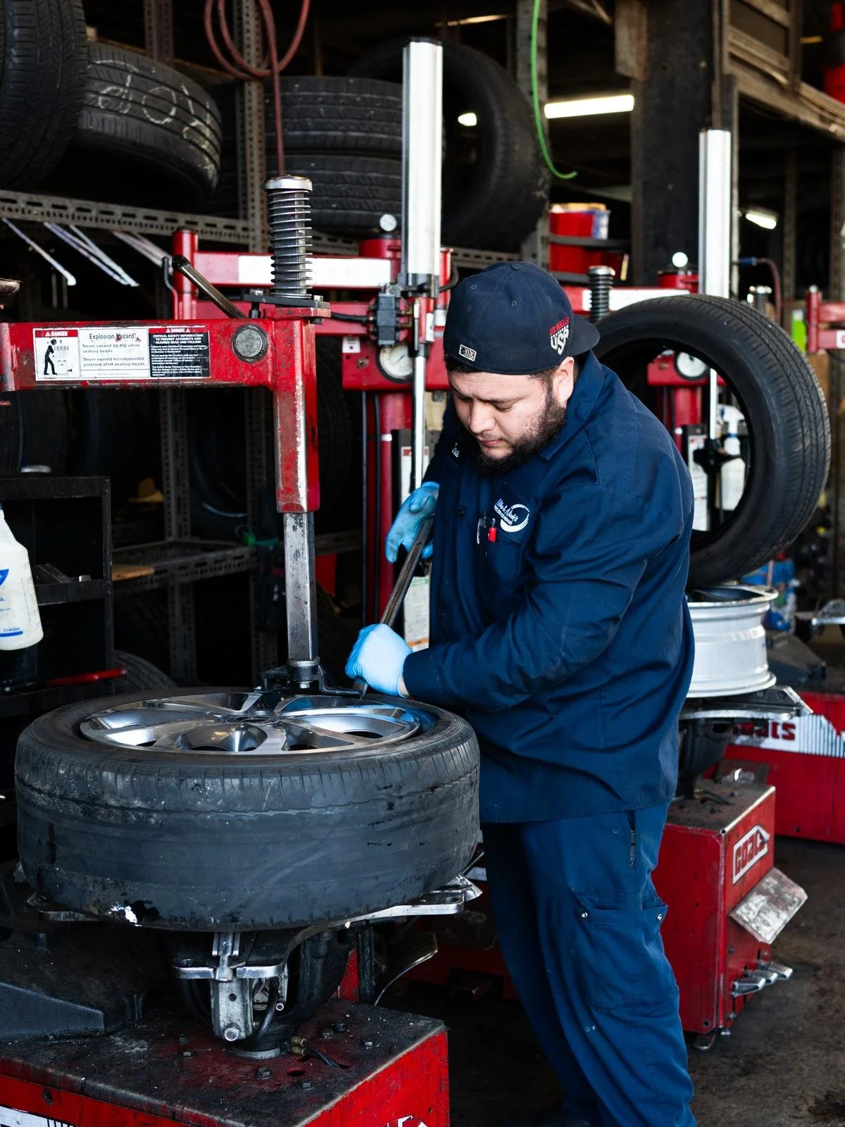 A mechanic working on a car tire using a tire balancing machine in a garage with shelves of tires and tools.