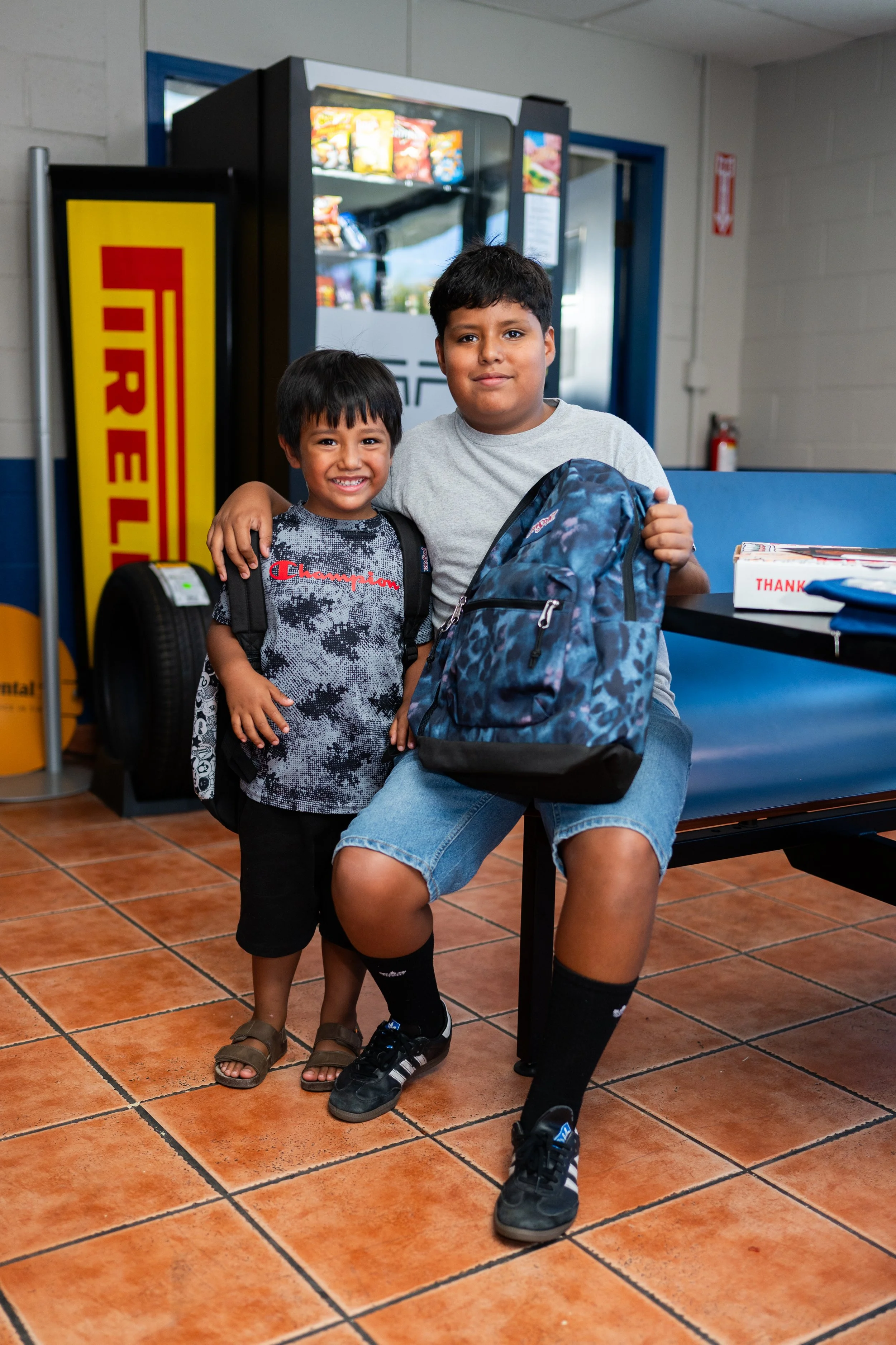 Two young boys stand inside a store near a vending machine. The older boy is sitting on a blue bench, holding a blue camo backpack, and has his arm around the younger boy, who is smiling. Both boys are dressed casually, and the store has a tiled floo