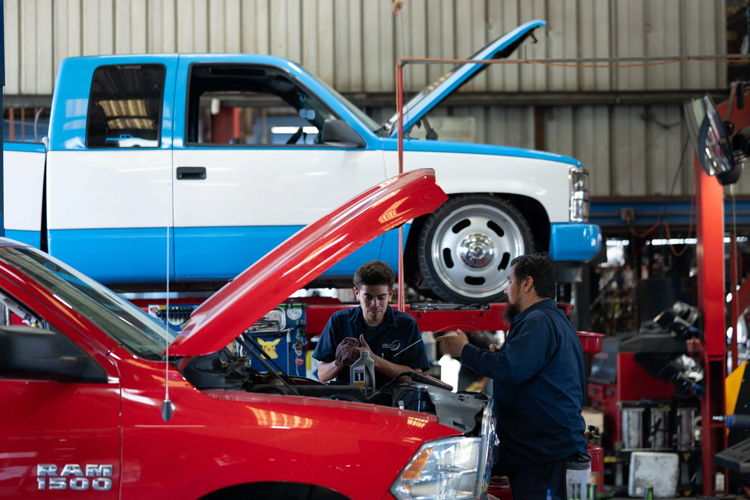 Automotive mechanics working on a red pickup truck inside an auto repair shop with a blue and white vehicle on lift in the background.