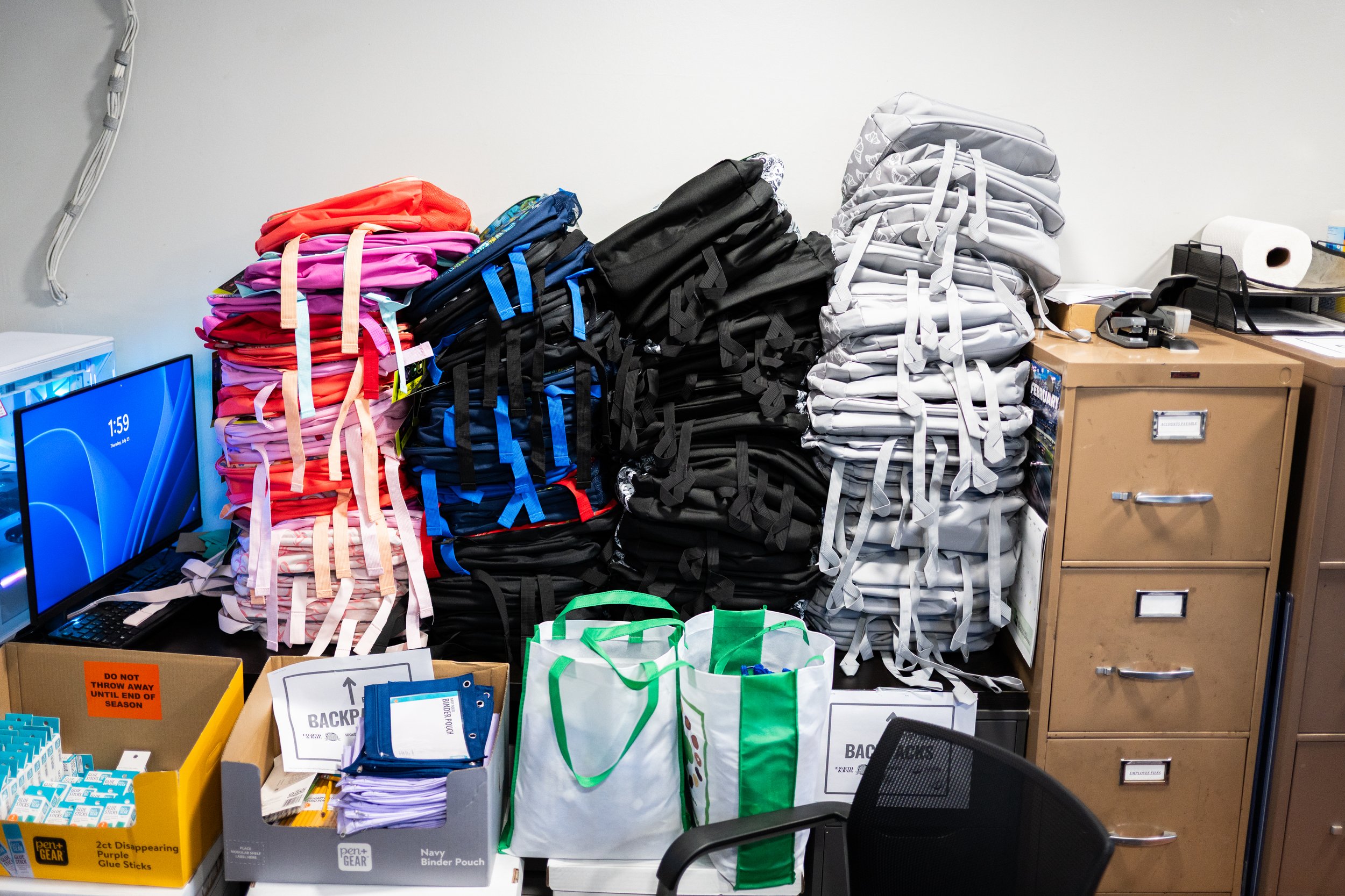 Stacks of backpacks and bags on a desk in an office or school supply room.