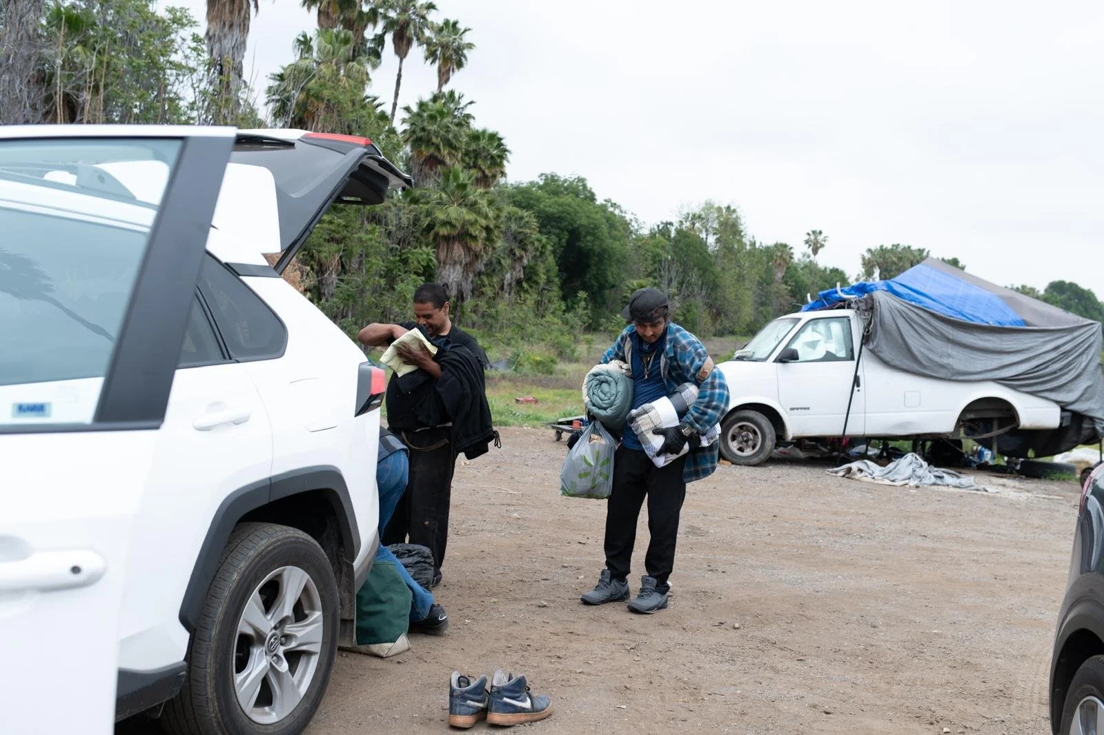 Two men are packing or unpacking cars on a dirt lot near a wooded area with palm trees. One man is loading items into a white SUV, and the other is holding a sleeping bag and other belongings. In the background, there is a white van covered with tarp