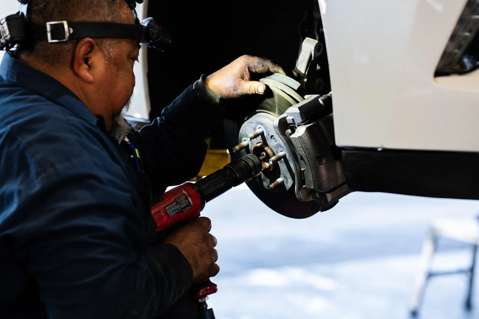 A mechanic working on a car's wheel hub, using a power drill, with the car raised on a lift.