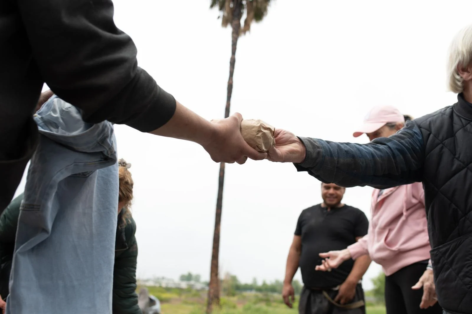 Two people are shaking hands; the person on the left is holding a wrapped item, and the person on the right has an outstretched hand. Several other people are in the background outdoors on a cloudy day.