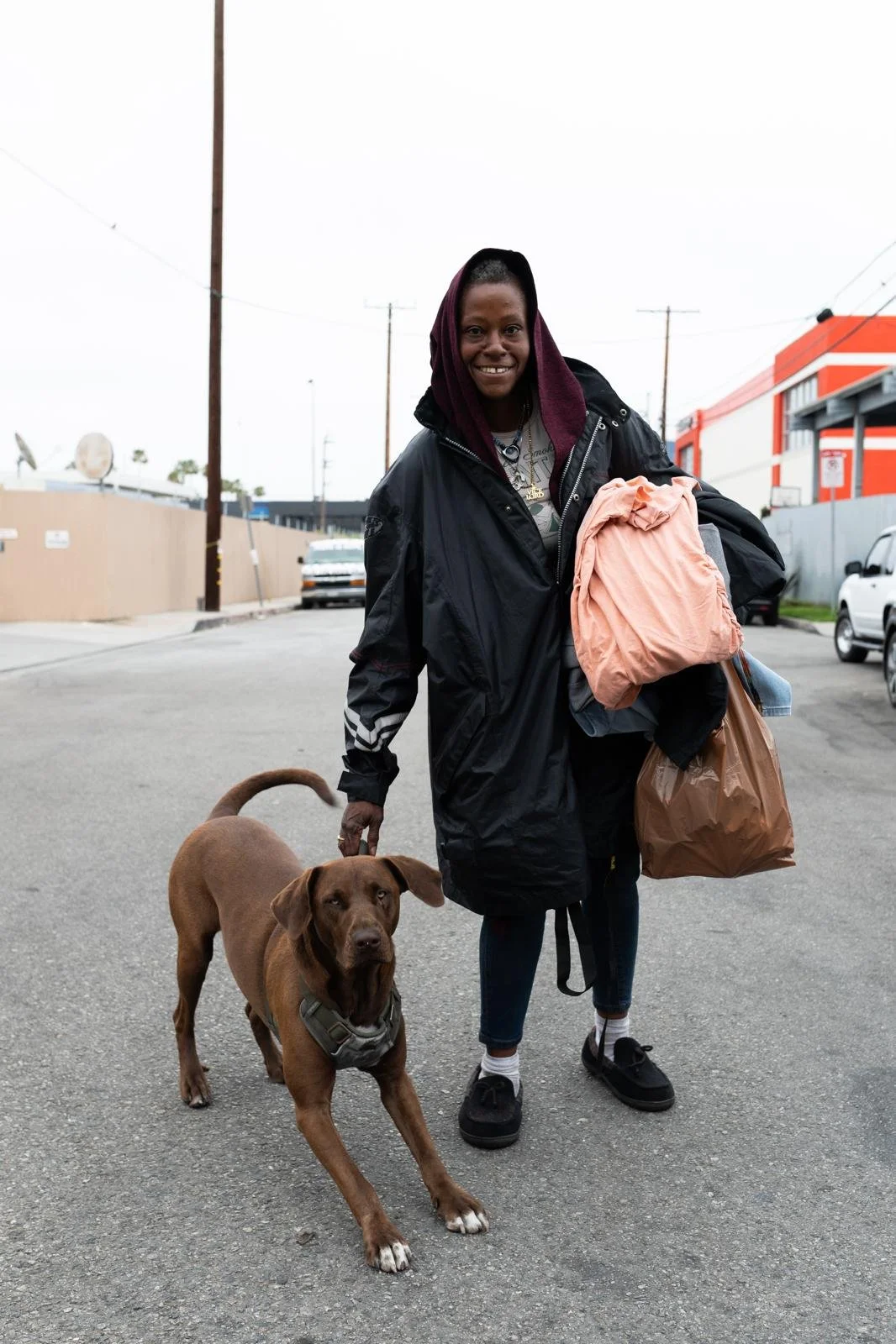 Woman smiling, holding a brown dog on a leash, carrying shopping bags, on a city street with parked cars and buildings.
