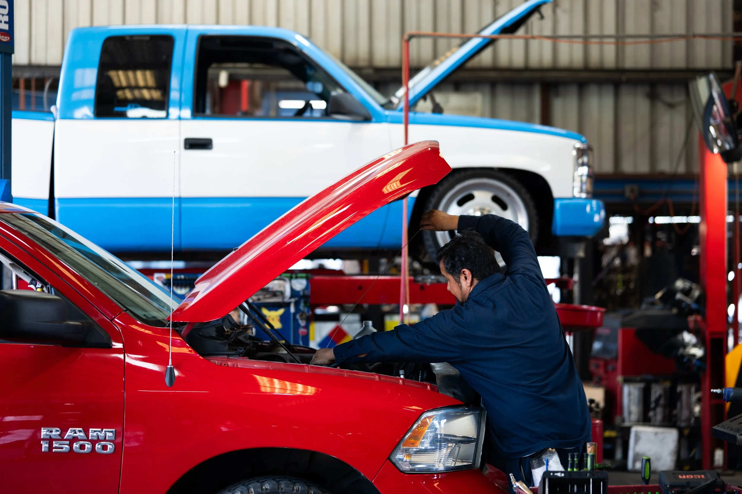 A mechanic working under the hood of a red Dodge Ram 1500 pickup truck in an auto repair shop, with another blue and white pickup truck on an overhead lift in the background.