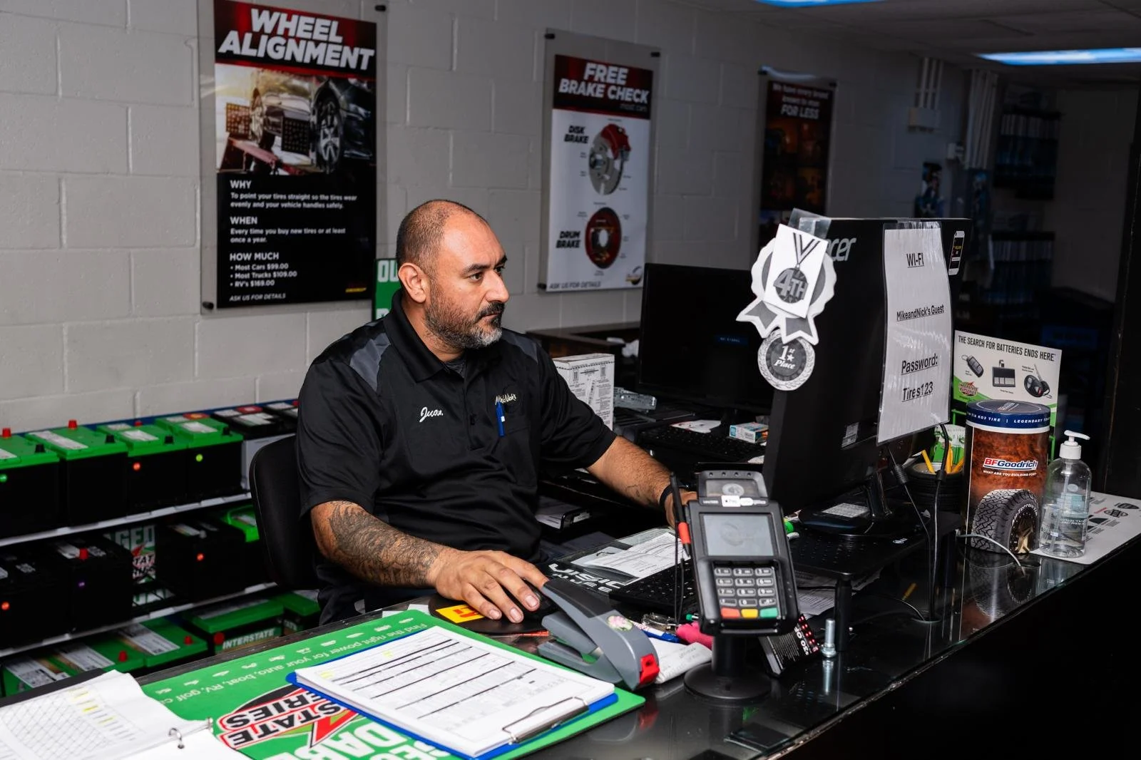 A man working at a desk in an auto service shop, surrounded by tools, signs, and equipment related to tire and wheel alignment services.