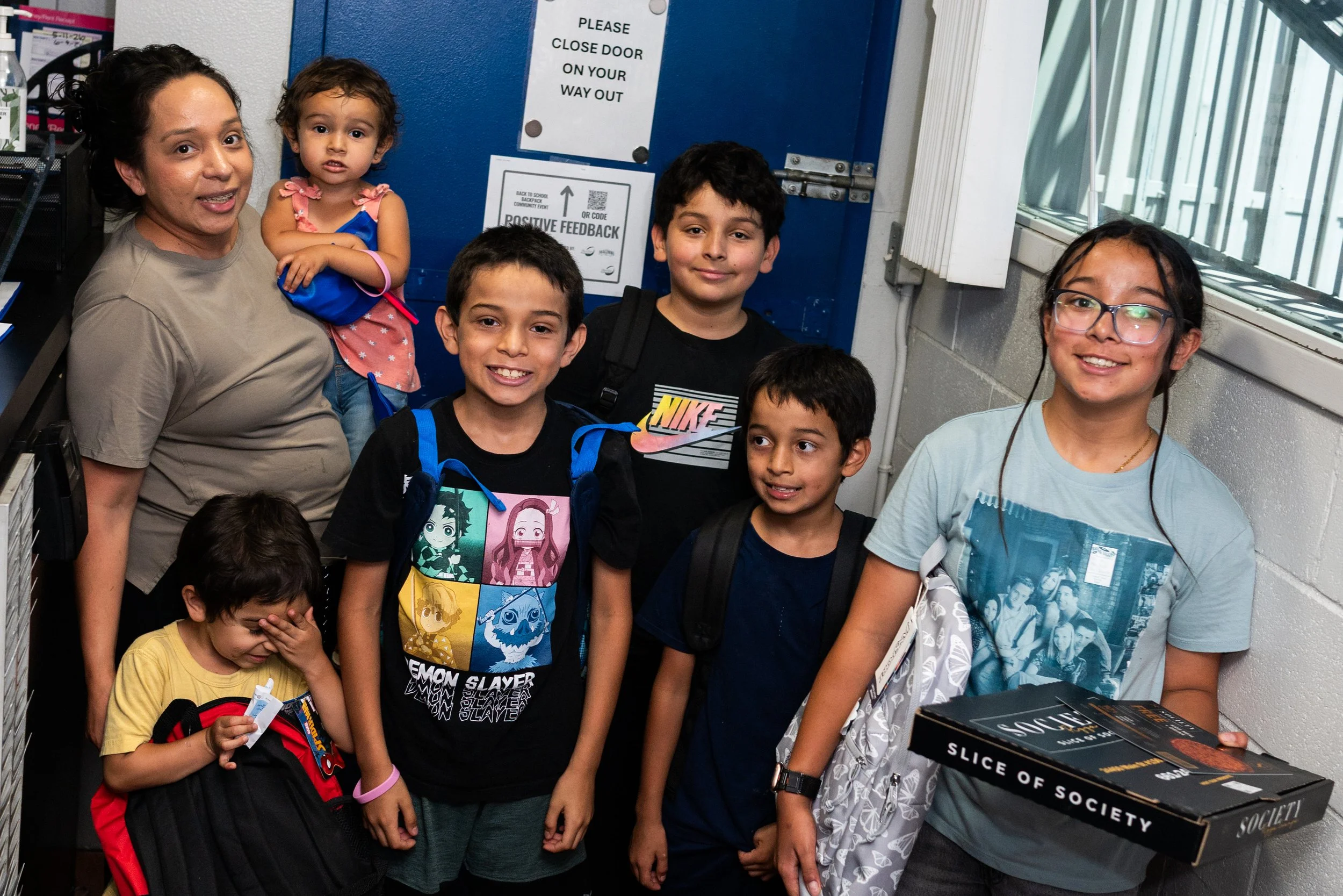 A group of children and a woman gathered inside a building near a door with signs, one reading 'PLEASE CLOSE DOOR ON YOUR WAY OUT' and another with a QR code and text about feedback. The children are smiling, with some wearing backpacks, and one girl