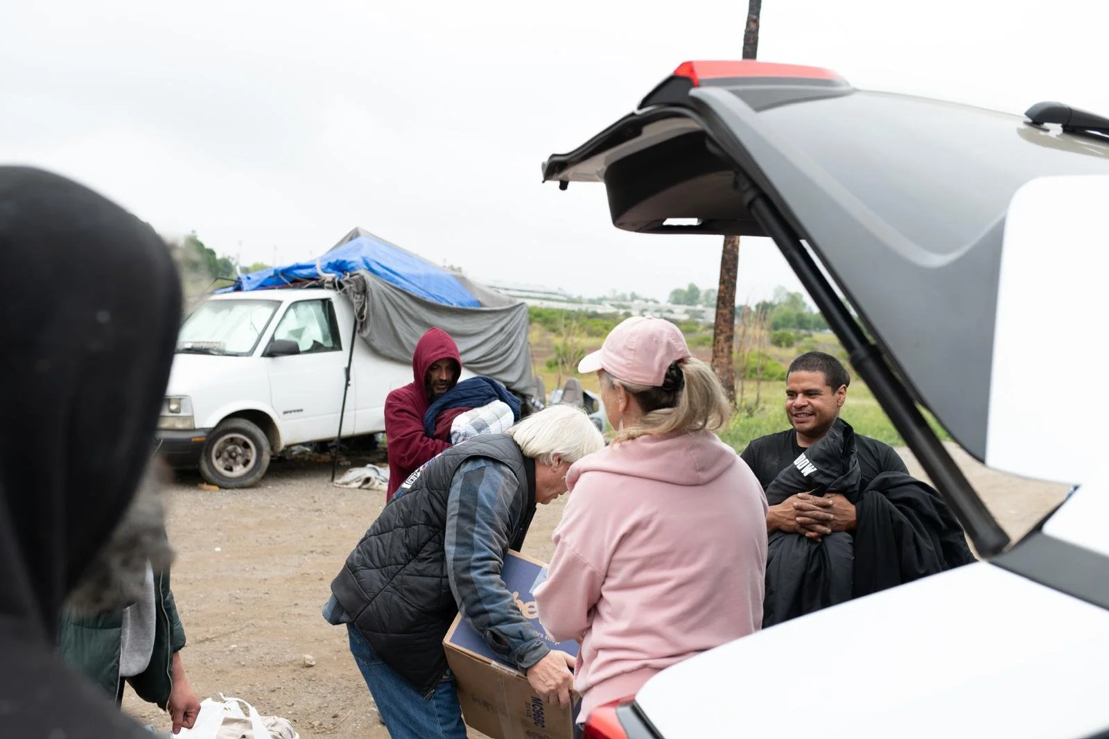 Group of people gathered outdoors by an open car trunk, with one woman handing a box to another woman, and a man sitting nearby smiling, in a rural or semi-rural setting with a white van and tent in the background.